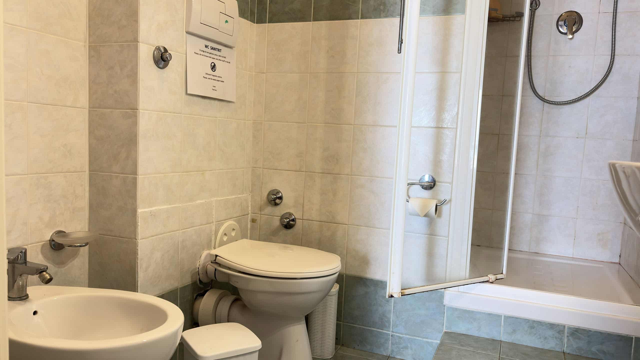 Spazio bagno accessibile con WC e doccia, rivestimenti in piastrelle beige, ambiente pulito e funzionale, ideale per soluzioni di arredo bagno moderne e pratiche.