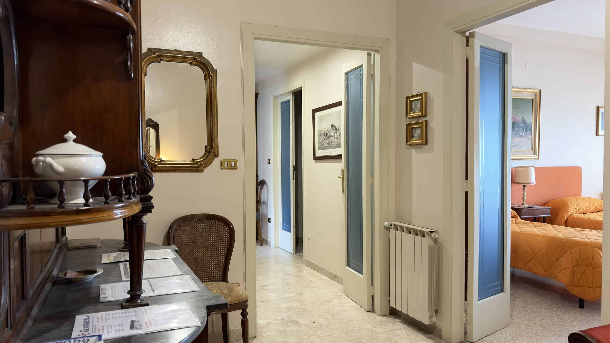 Elegante ingresso di casa con pareti chiare, porta aperta che conduce a un soggiorno luminoso con divano arancione e decorazioni moderne.