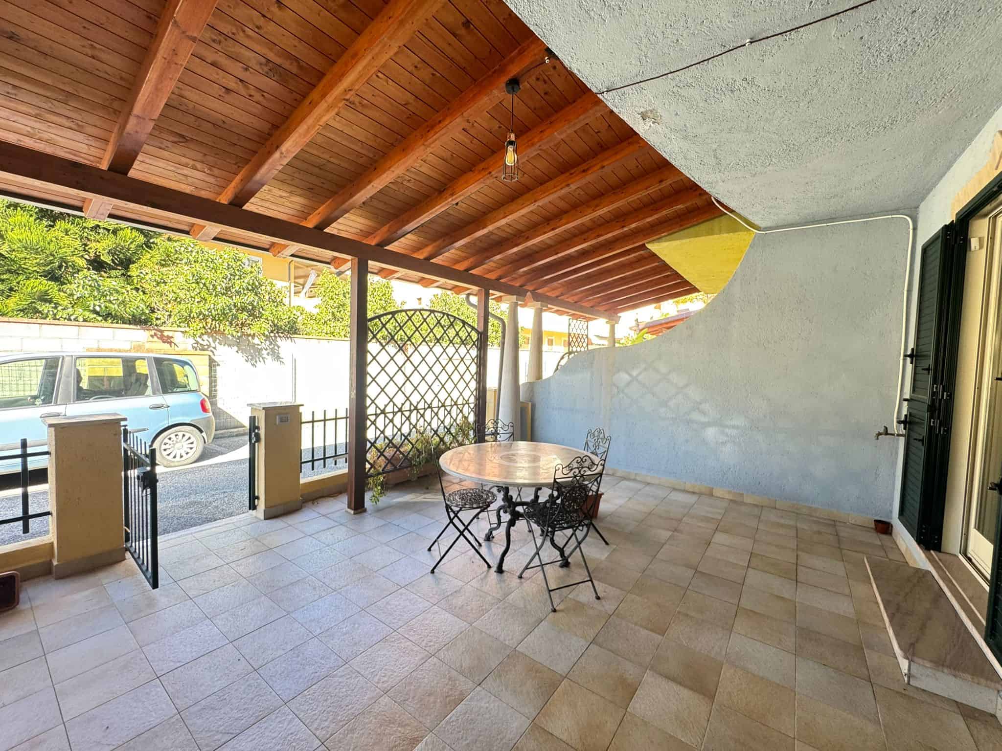 Ampia terrazza coperta con arredi in ferro battuto, vista su strada e giardino, ideale per relax e pranzi all'aperto, in zona residenziale tranquilla vicino a servizi e spazi verdi.