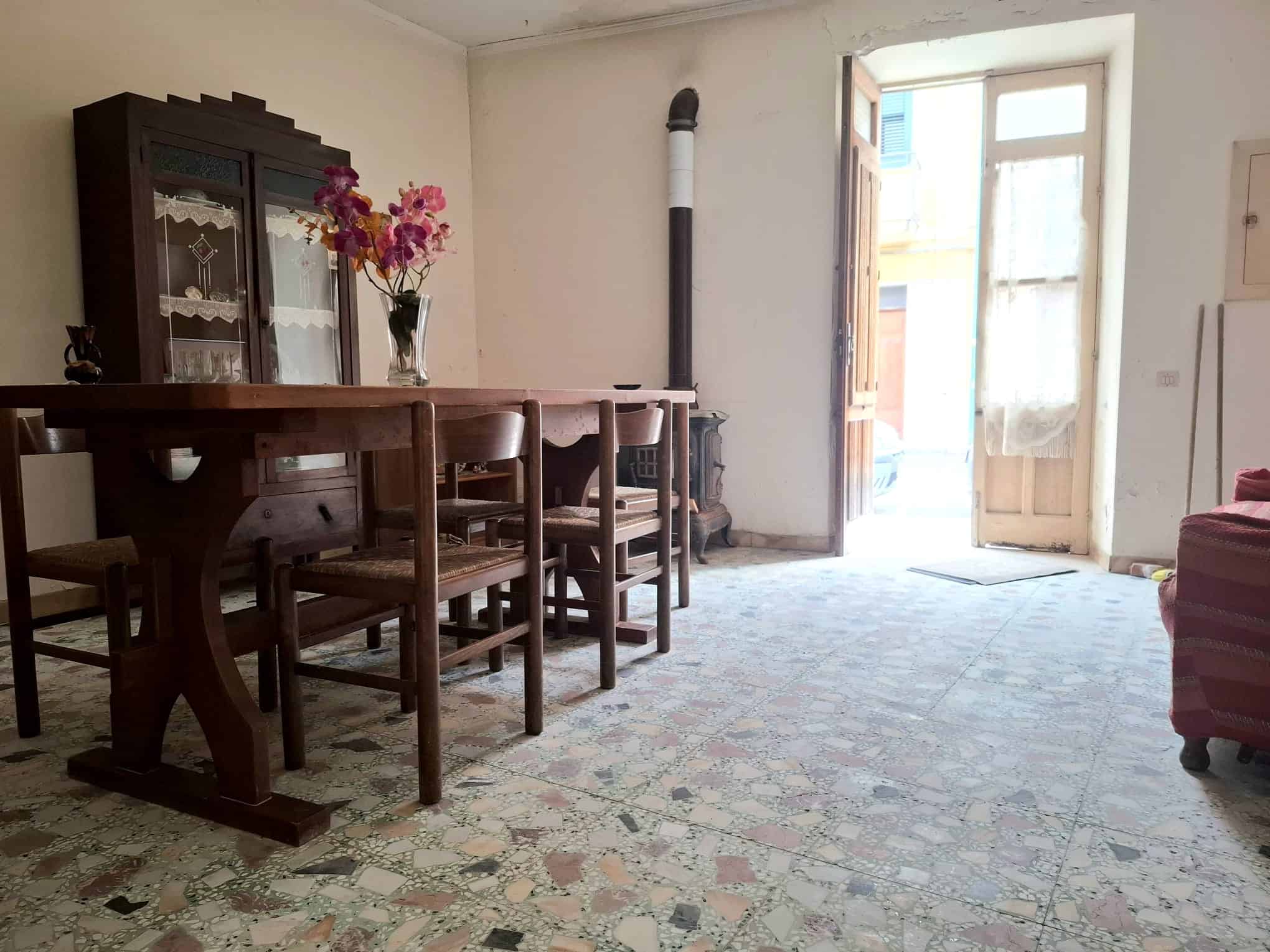 Panca di legno con sedie in una stanza luminosa e spaziosa, porta finestra che apre su un balcone, arredamento semplice e atmosfera accogliente, ideale per soggiorno o sala da pranzo.