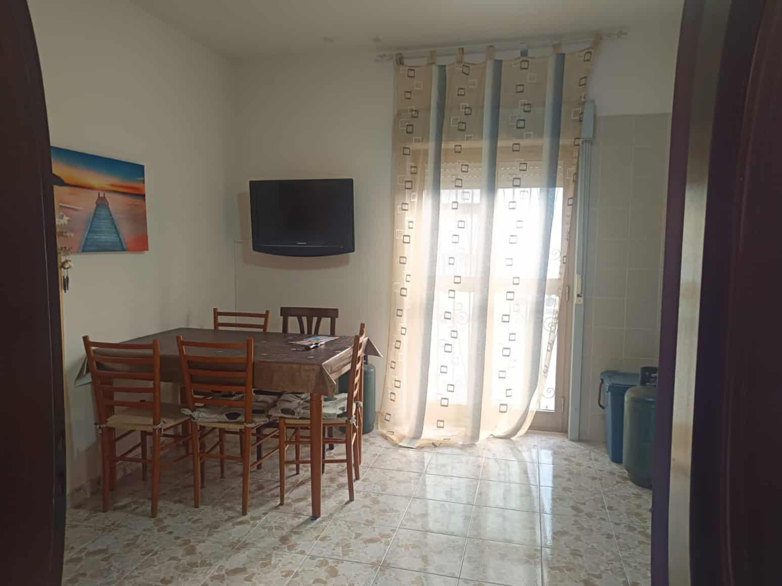 Interno di un soggiorno con tavolo da pranzo, televisore e balcone con tende leggere, arredamento semplice e luminoso, ideale per soggiorni confortevoli.