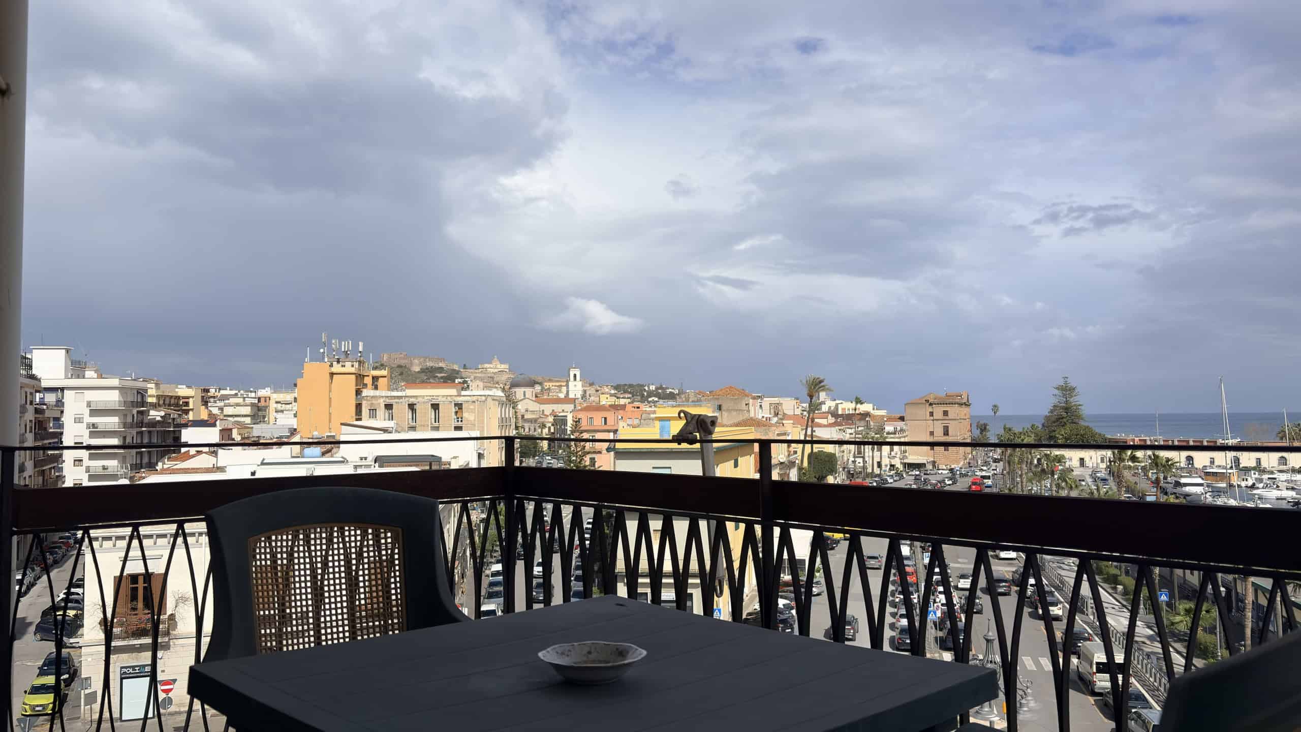 Balcone con vista sulla città e cielo nuvoloso, arredato con tavolo nero e sedia in plastica, ideale per relax e momenti all'aperto.