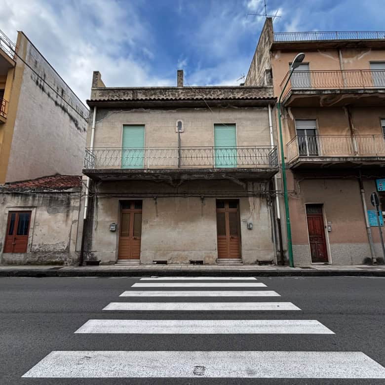 Edificio storico in centro città con facciata in pietra e balconi in ferro battuto, situato lungo una strada trafficata con strisce pedonali in primo piano, rappresenta un esempio di architettura urba.