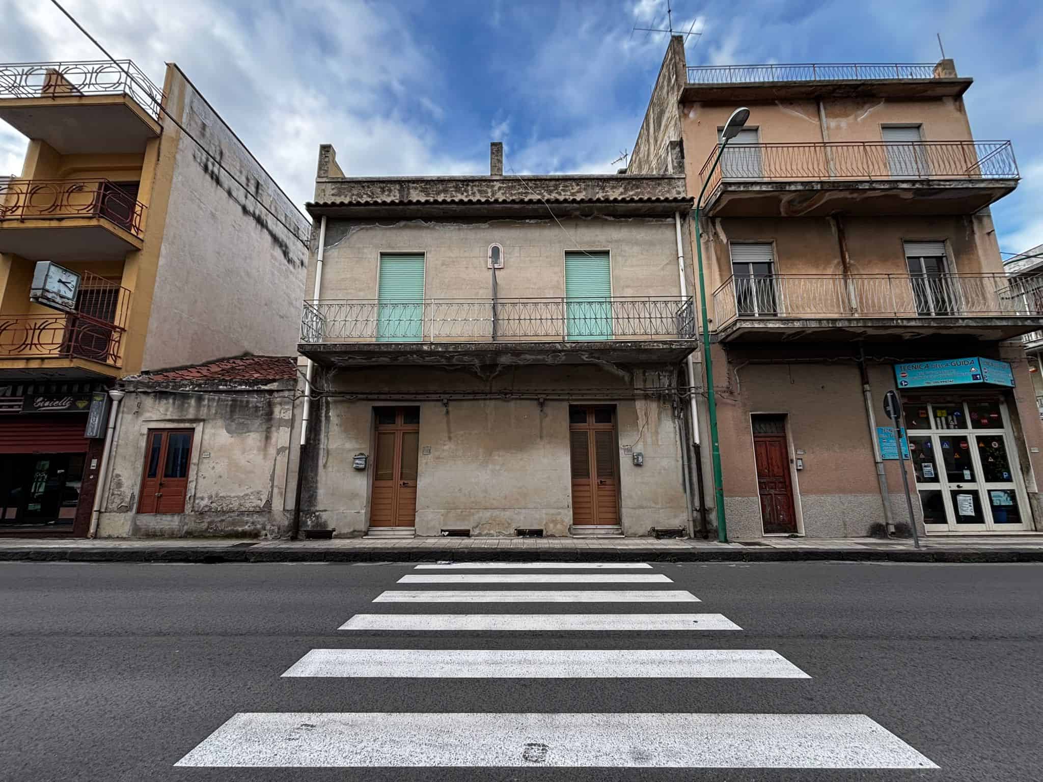 Edificio storico in centro città con facciata in pietra e balconi in ferro battuto, situato lungo una strada trafficata con strisce pedonali in primo piano, rappresenta un esempio di architettura urba.
