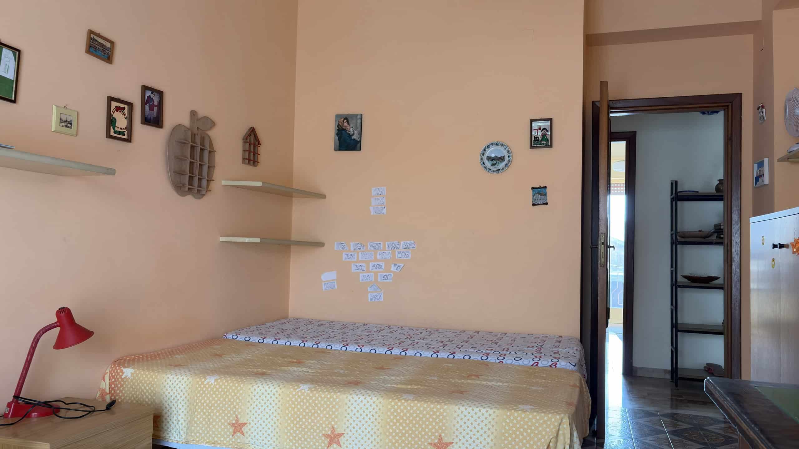 Elegante camera da letto con pareti color pesca, decorata con quadri e orologi, letto matrimoniale con copriletto beige con stelle, lampada da tavolo rossa e scaffali a parete.