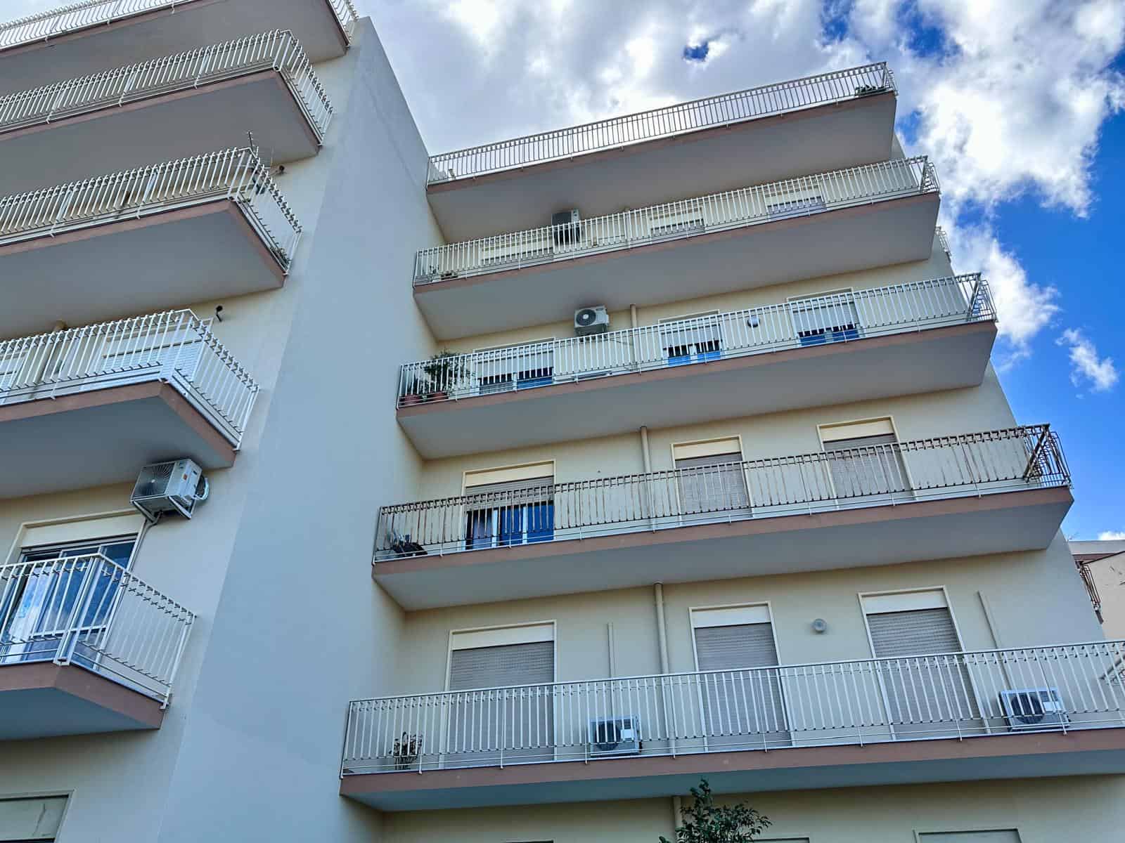 Balcone di un edificio residenziale moderno con ringhiere bianche e aria condizionata, sotto un cielo azzurro con nuvole bianche, rappresenta un appartamento in affitto o vendita a NextCasa.