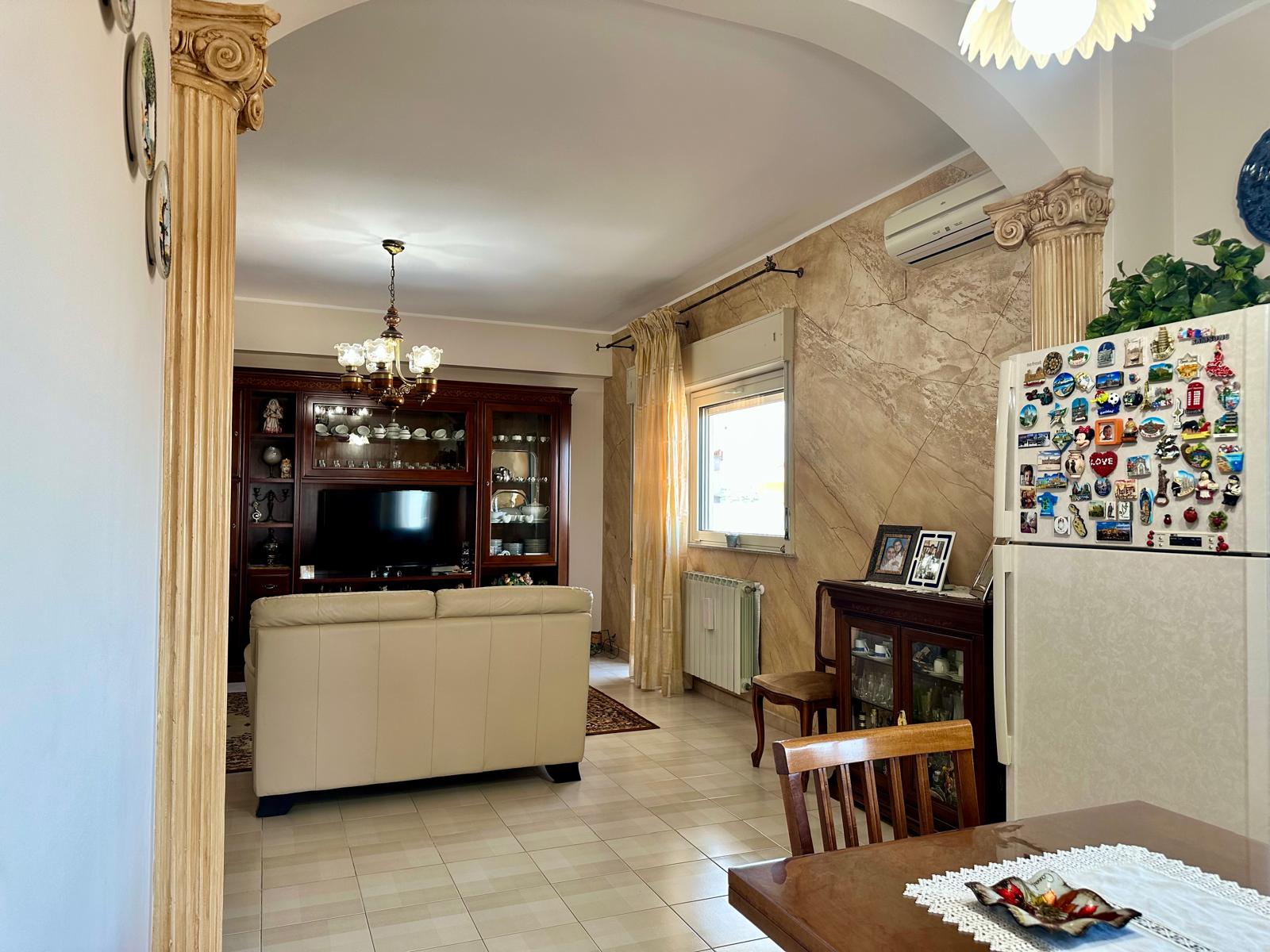Ampia zona living con divano beige, mobili eleganti e decorazioni classiche, luminosa con finestra e aria condizionata, ideale per soggiorni confortevoli e accoglienti in un ambiente raffinato.