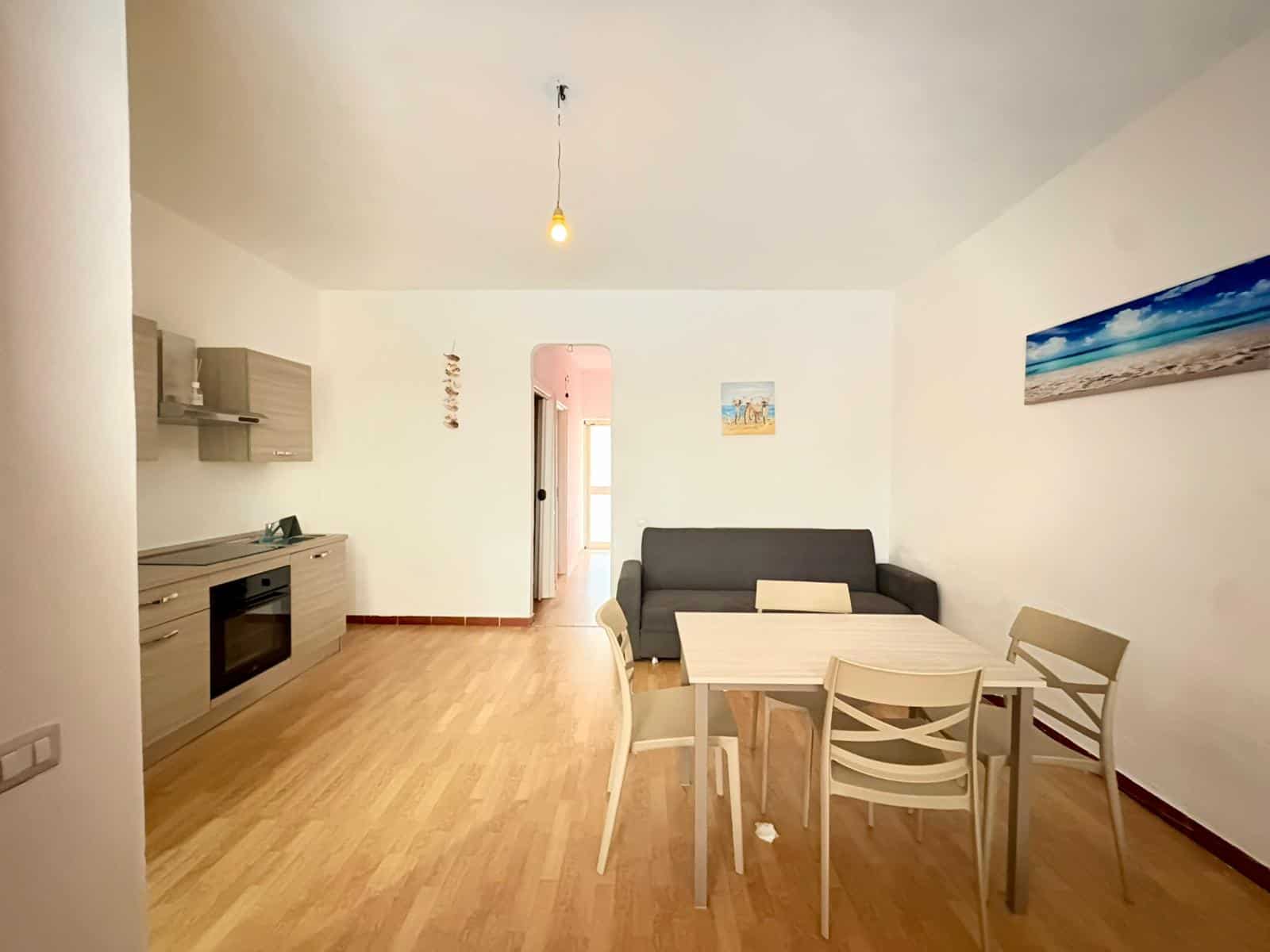 Ampio soggiorno open space con cucina moderna, zona pranzo e area living, arredamento minimalista, pavimento in legno chiaro, pareti bianche, decorazioni colorate, illuminazione naturale e lampada a s.