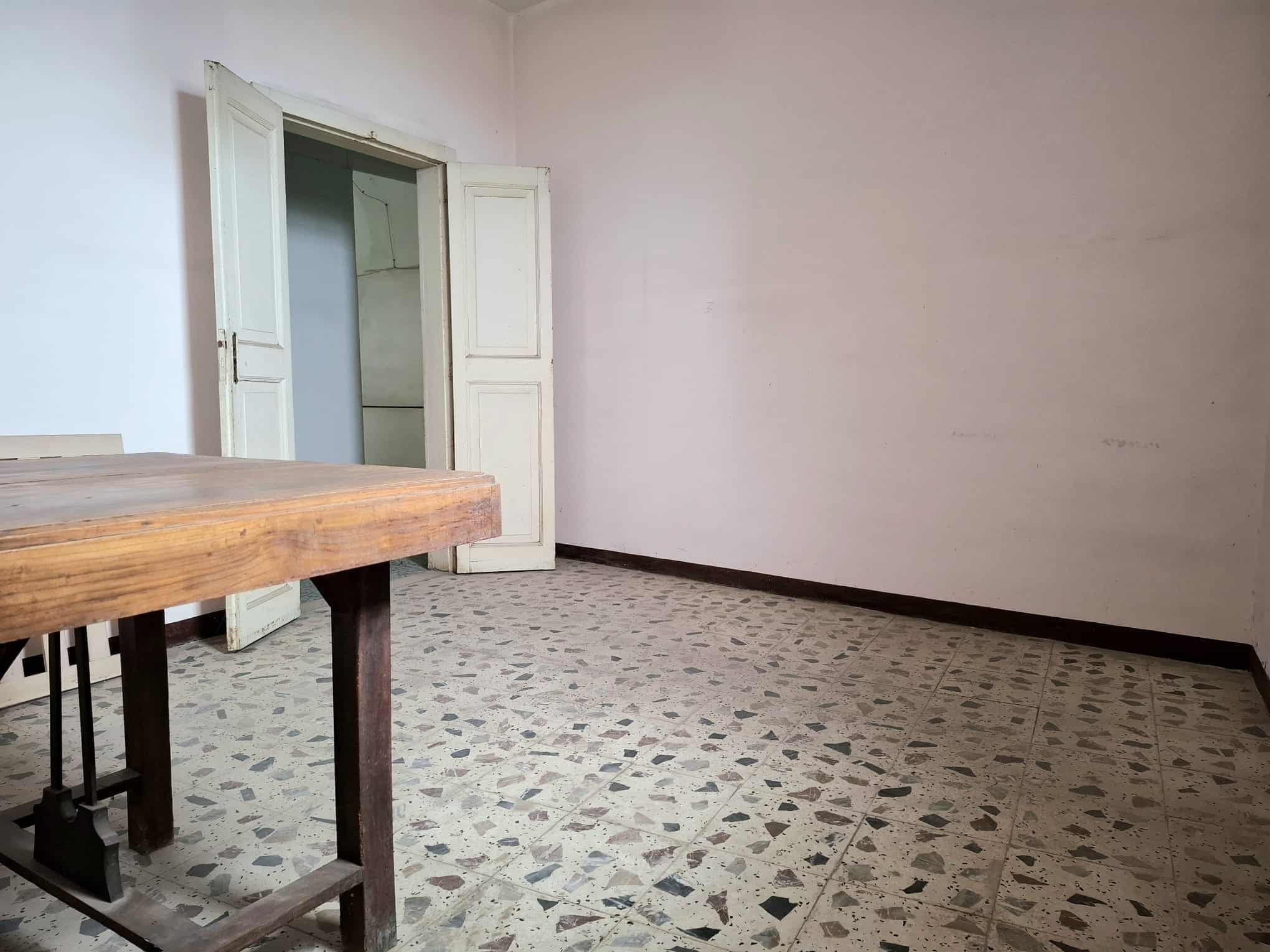 Una stanza vuota con pareti bianche, pavimento in piastrelle e una porta aperta che conduce a un'altra stanza, con un tavolo in legno nella parte sinistra.