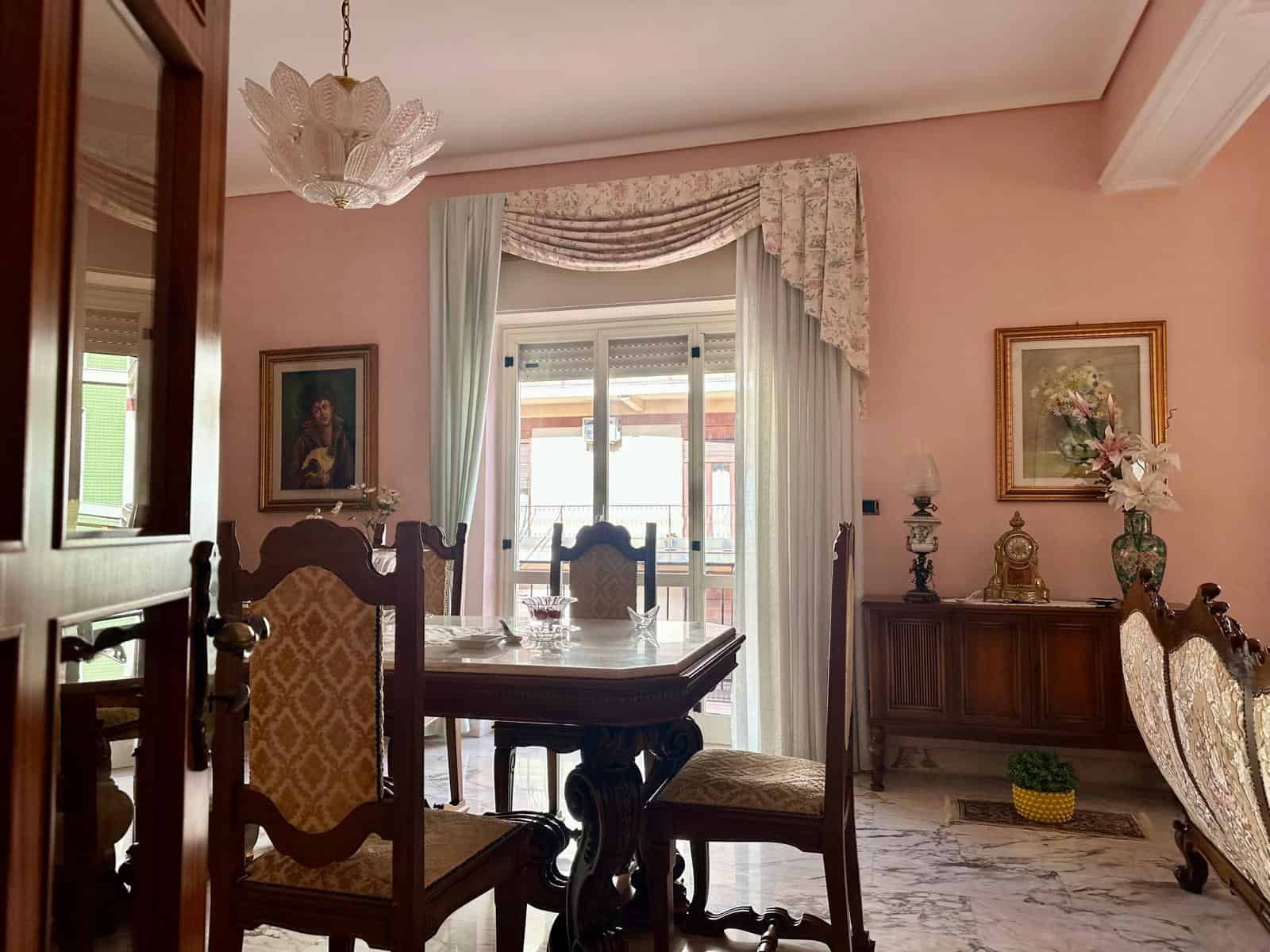Elegante sala da pranzo con mobili in legno, tende leggere e decorazioni floreali, ambiente accogliente e raffinato.