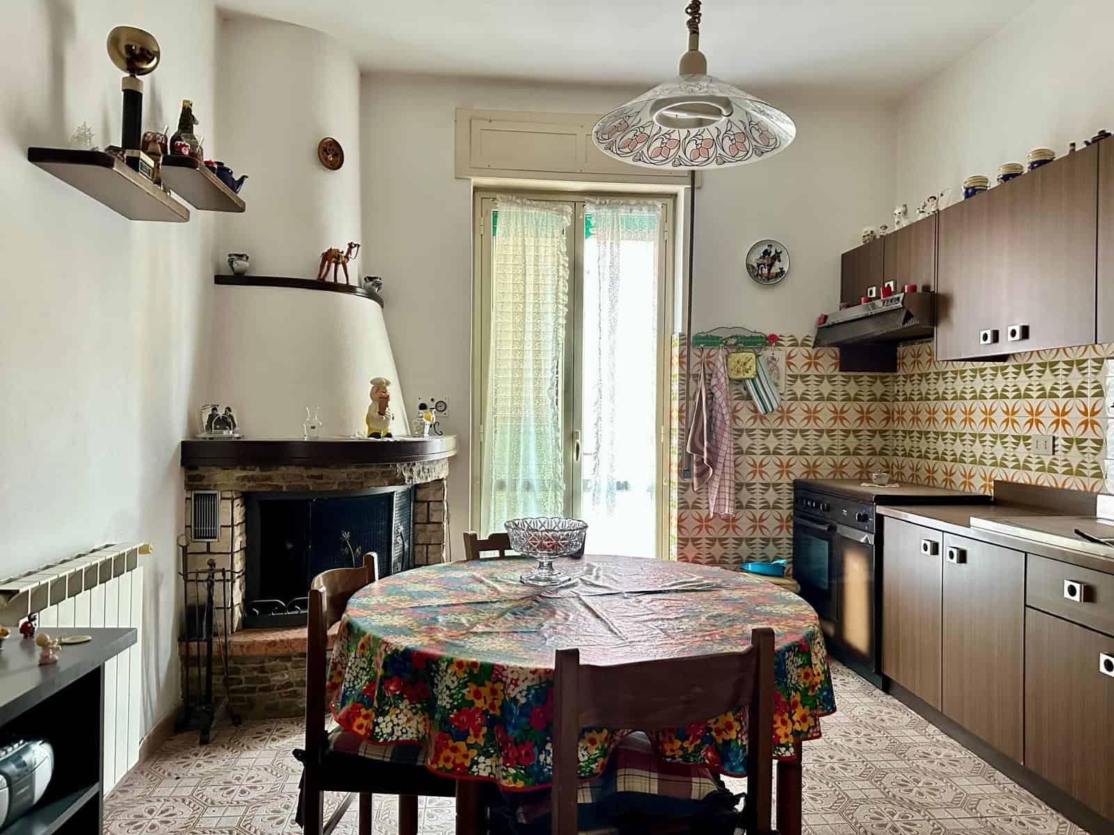 Pannello di cucina e soggiorno con camino, tavolo rotondo con tovaglia floreale, mobili in legno chiaro, pareti decorate con piastrelle colorate, finestra con tende leggere, atmosfera accogliente e tr.