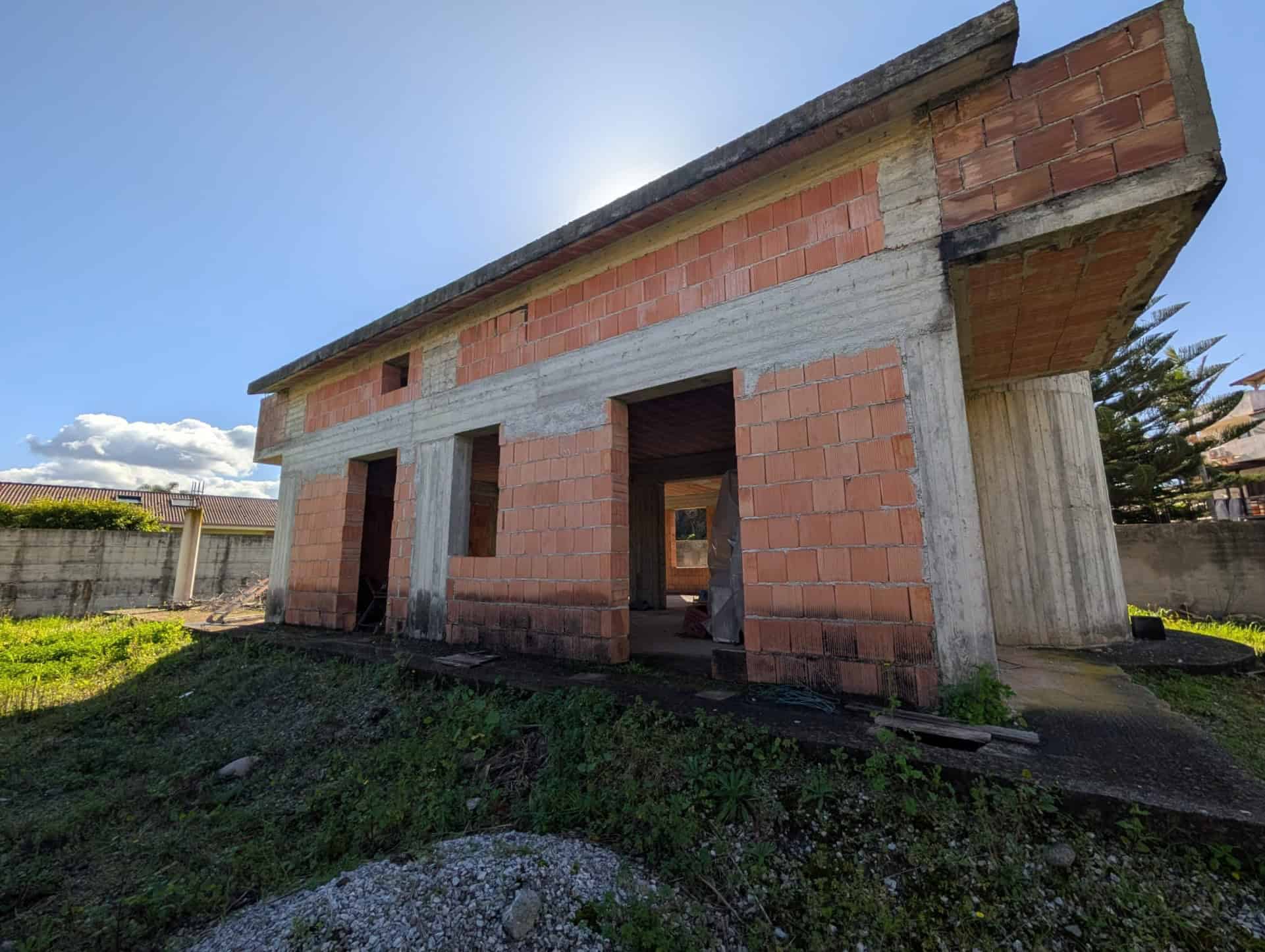 Pannello di casa in costruzione con struttura in mattoni e cemento, pronto per finiture e rifiniture esterne, in un'area residenziale soleggiata e verdeggiante.