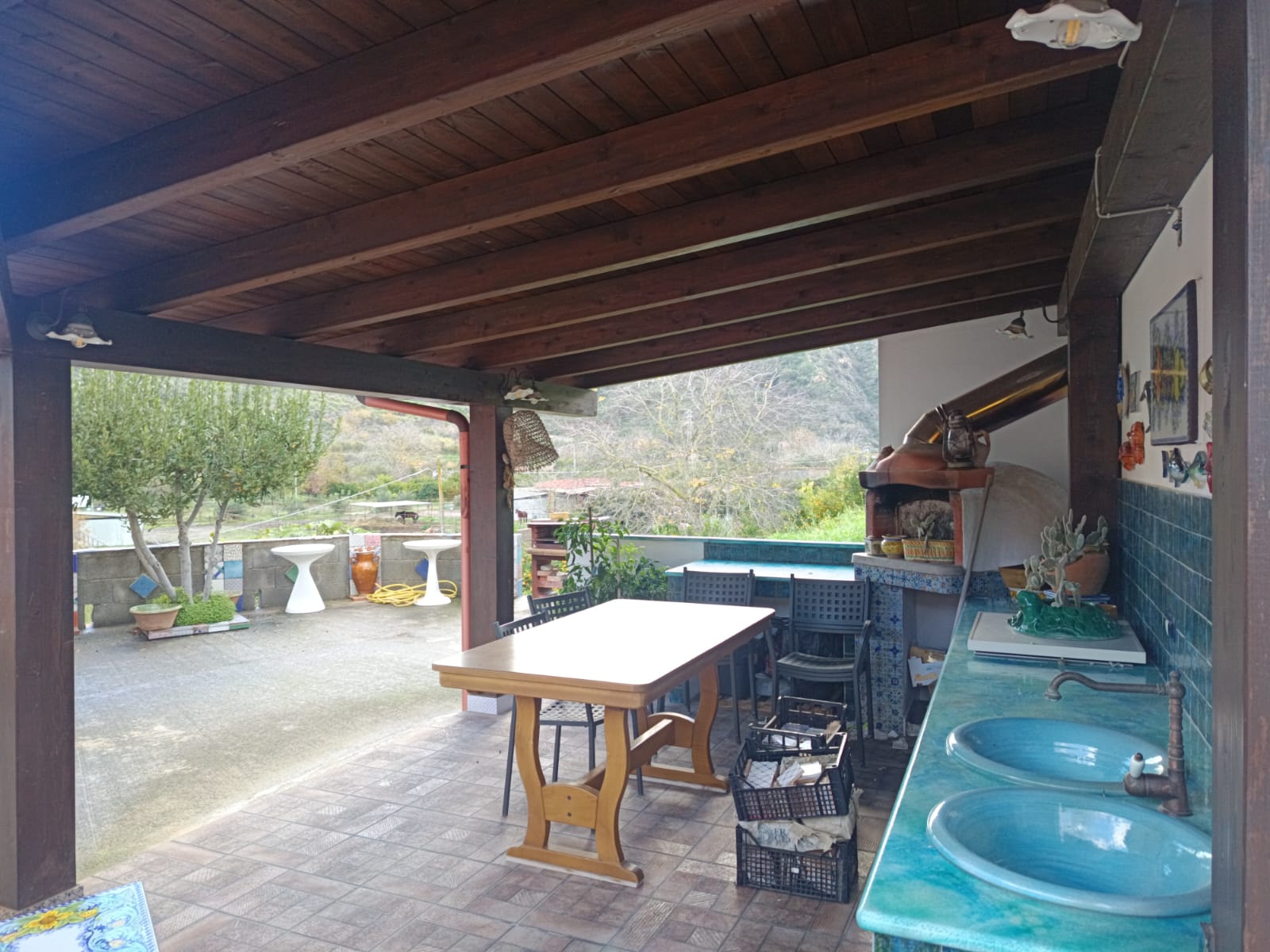Aggiungi un'immagine di una veranda esterna con tavolo e sedie, vista su un giardino con alberi e panchine, ideale per relax e convivialità all'aperto.