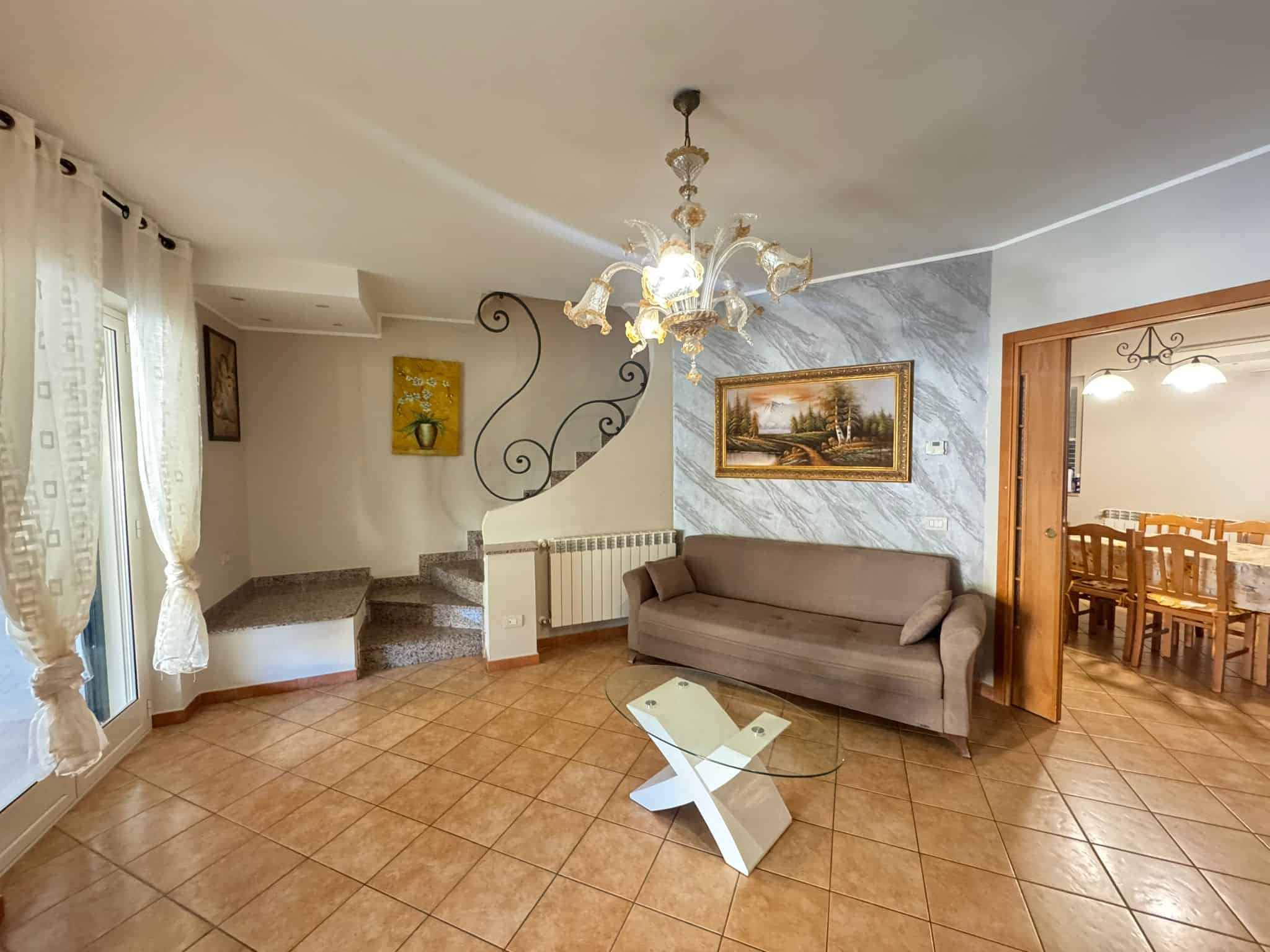 Ampio soggiorno con divano, lampadario elegante e pareti decorate, ideale per relax e incontri familiari, con accesso a una terrazza luminosa e spaziosa.