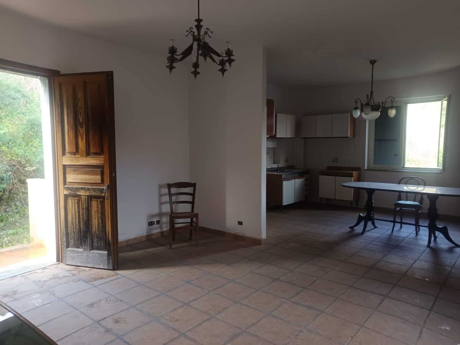 Porte in legno aperta che conduce a un soggiorno spazioso con cucina a vista, pavimento in piastrelle e illuminazione naturale da grandi finestre, ideale per ambienti di vita accoglienti e funzionali.