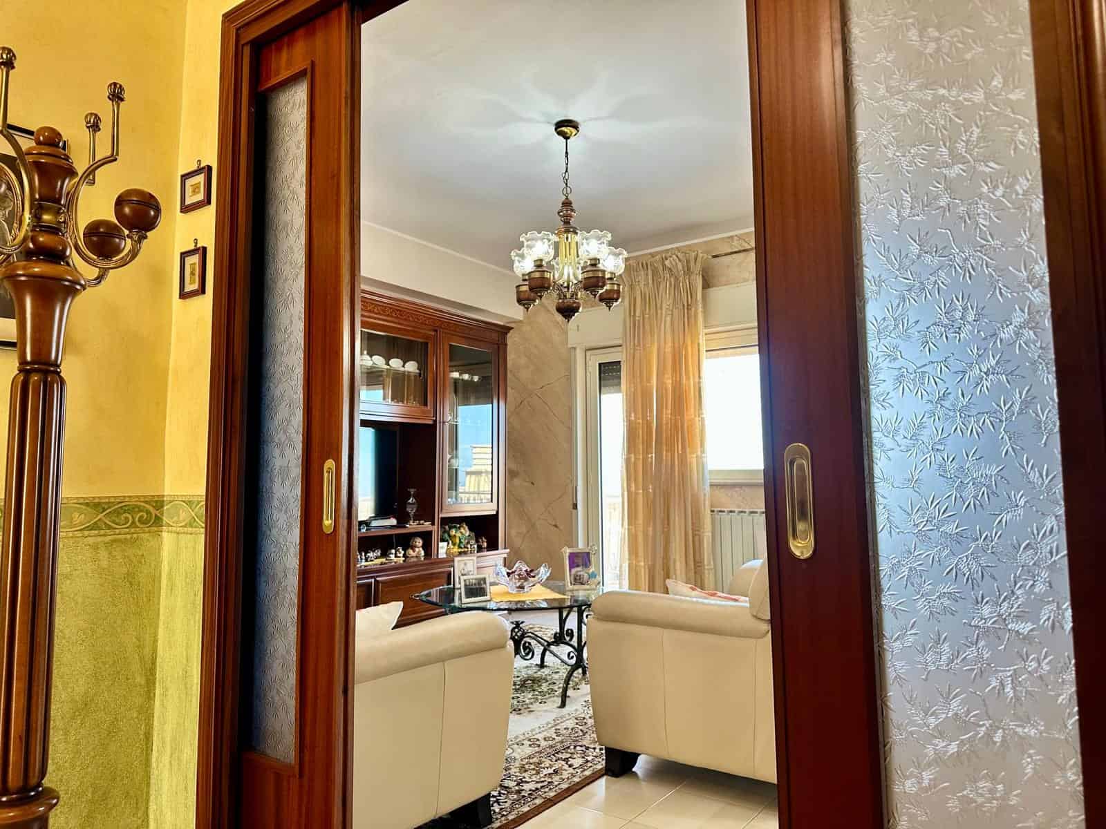 Elegante soggiorno con divani bianchi, camino in legno e grande finestra con tende leggere, illuminato da lampadario classico, ideale per vivere momenti di relax e convivialità.