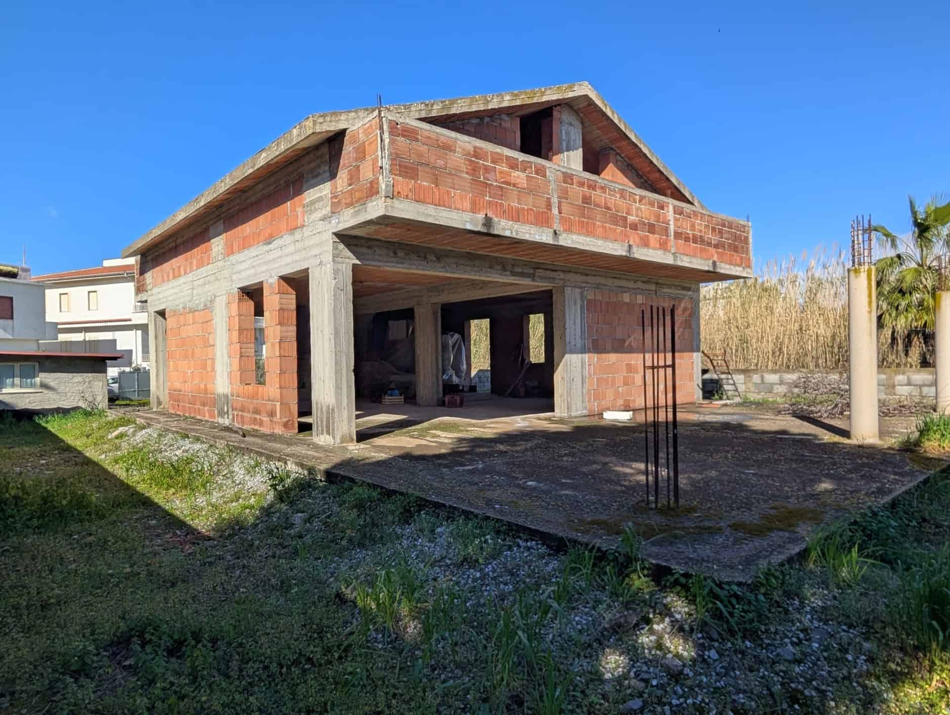 Pannello di casa in costruzione con struttura in cemento e mattoni rossi, pronto per essere completato, situata in un'area verde soleggiata con altre abitazioni nelle vicinanze.