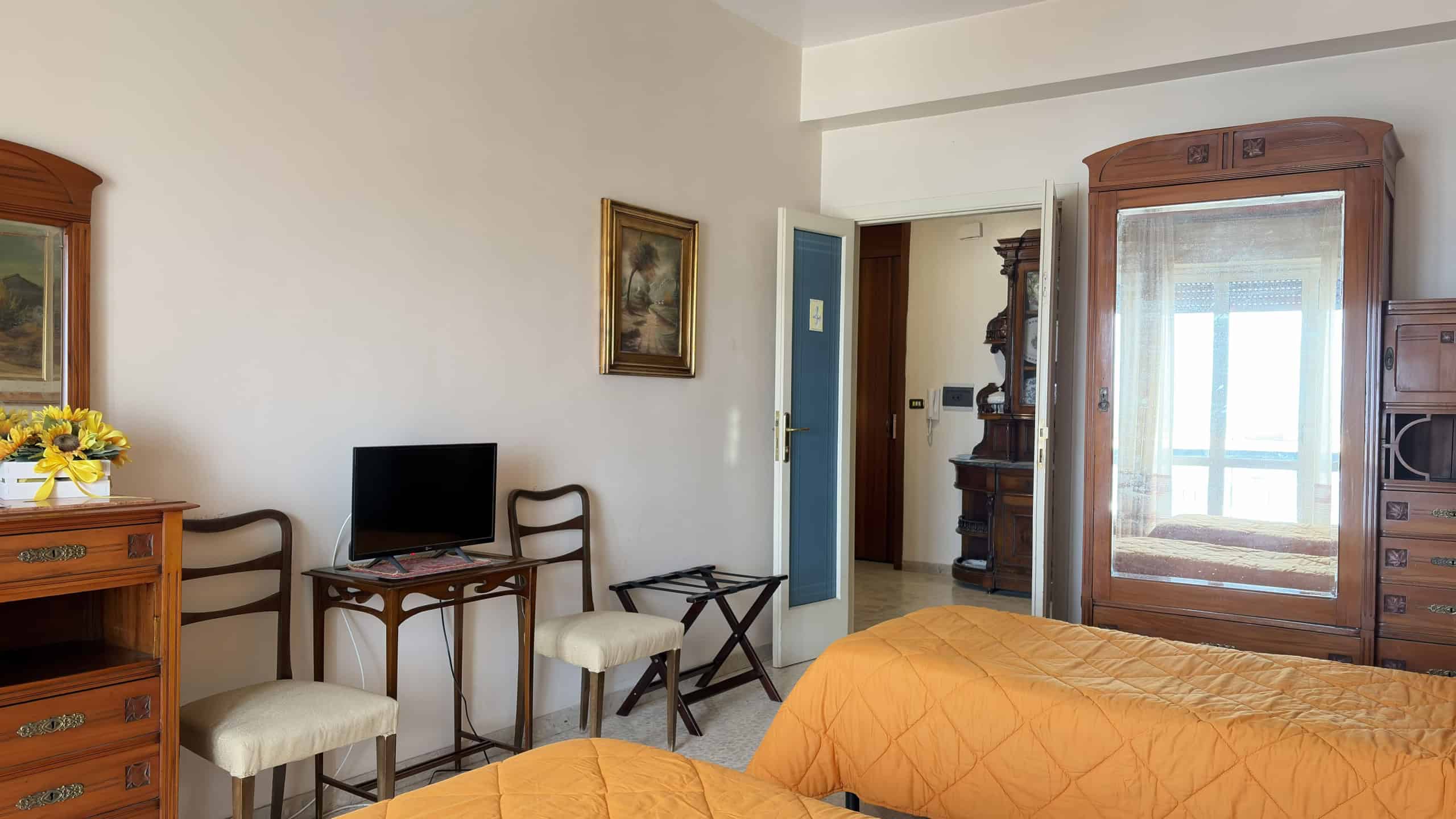 Riproduzione di una camera da letto classica con mobili in legno, letto con copertura arancione, armadio antico e sedie in stile tradizionale, ambiente luminoso e accogliente.