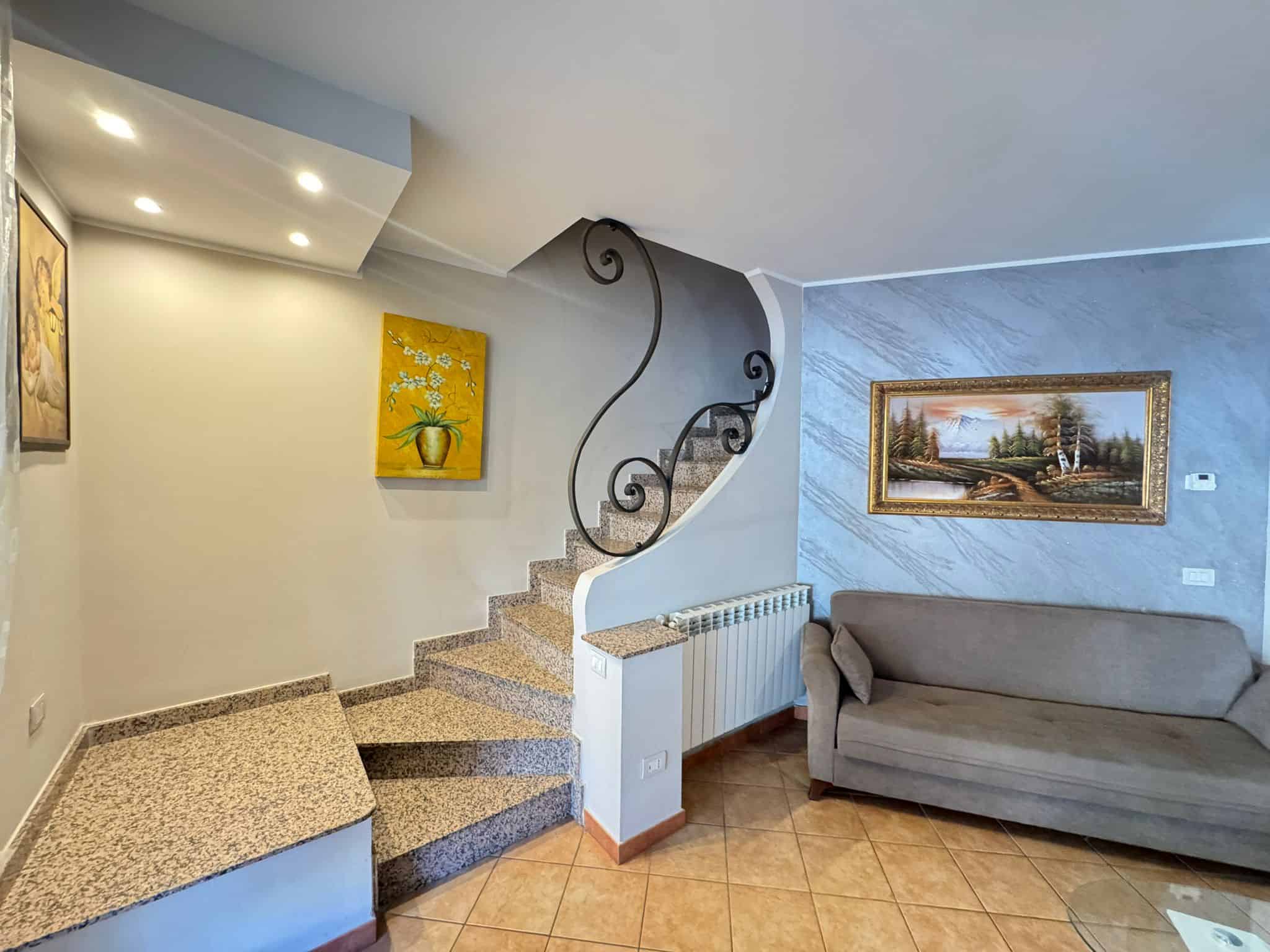 Un ingresso elegante con scala a chiocciola in marmo e ringhiera in ferro battuto, arredato con un divano grigio e un quadro paesaggistico alle pareti, in un ambiente luminoso e accogliente.