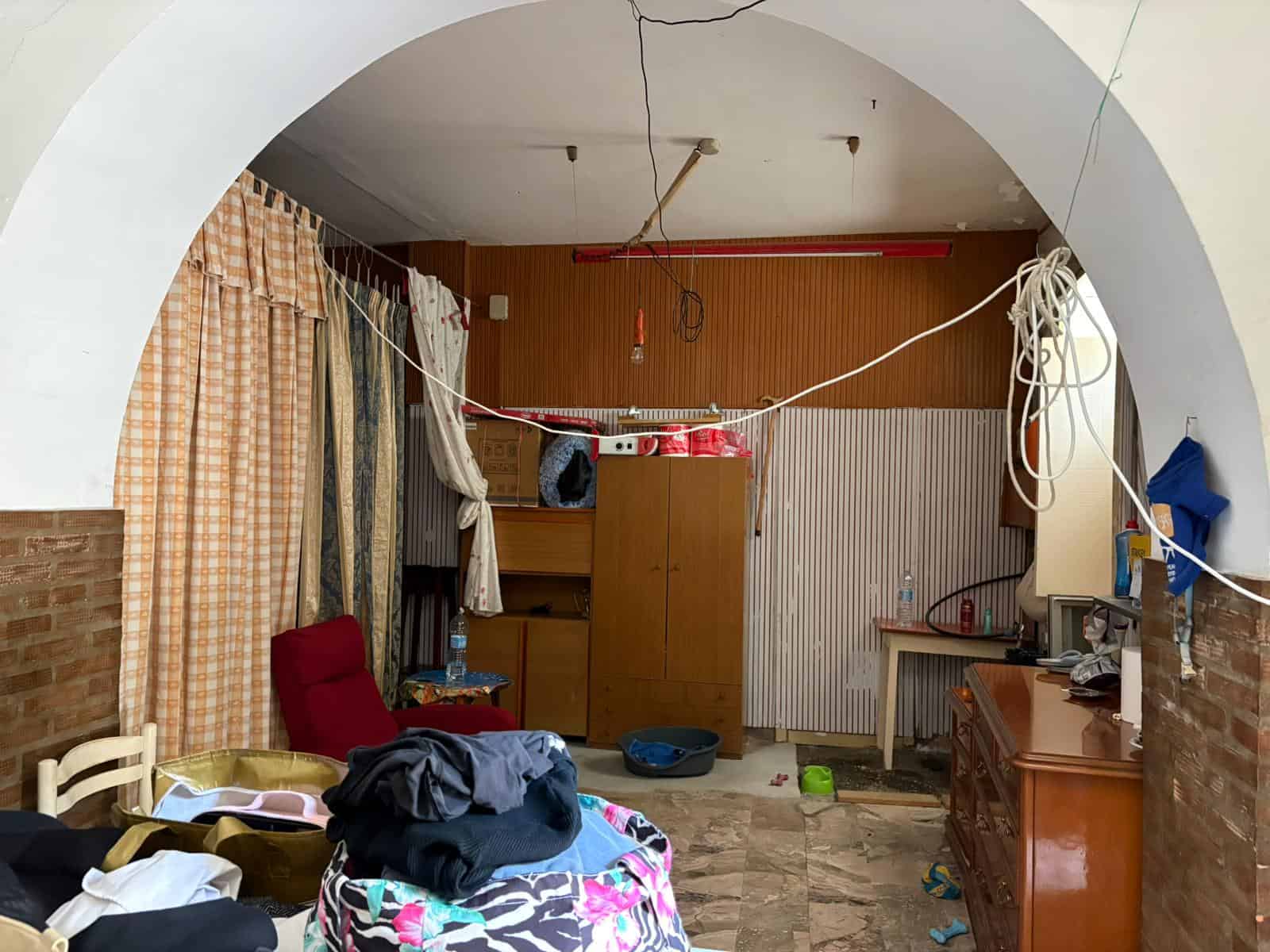 Senza titolo, interno di una casa disordinata con mobili e oggetti sparsi, pareti in mattoni e soffitto con fili pendenti, ambiente poco curato e trasandato.