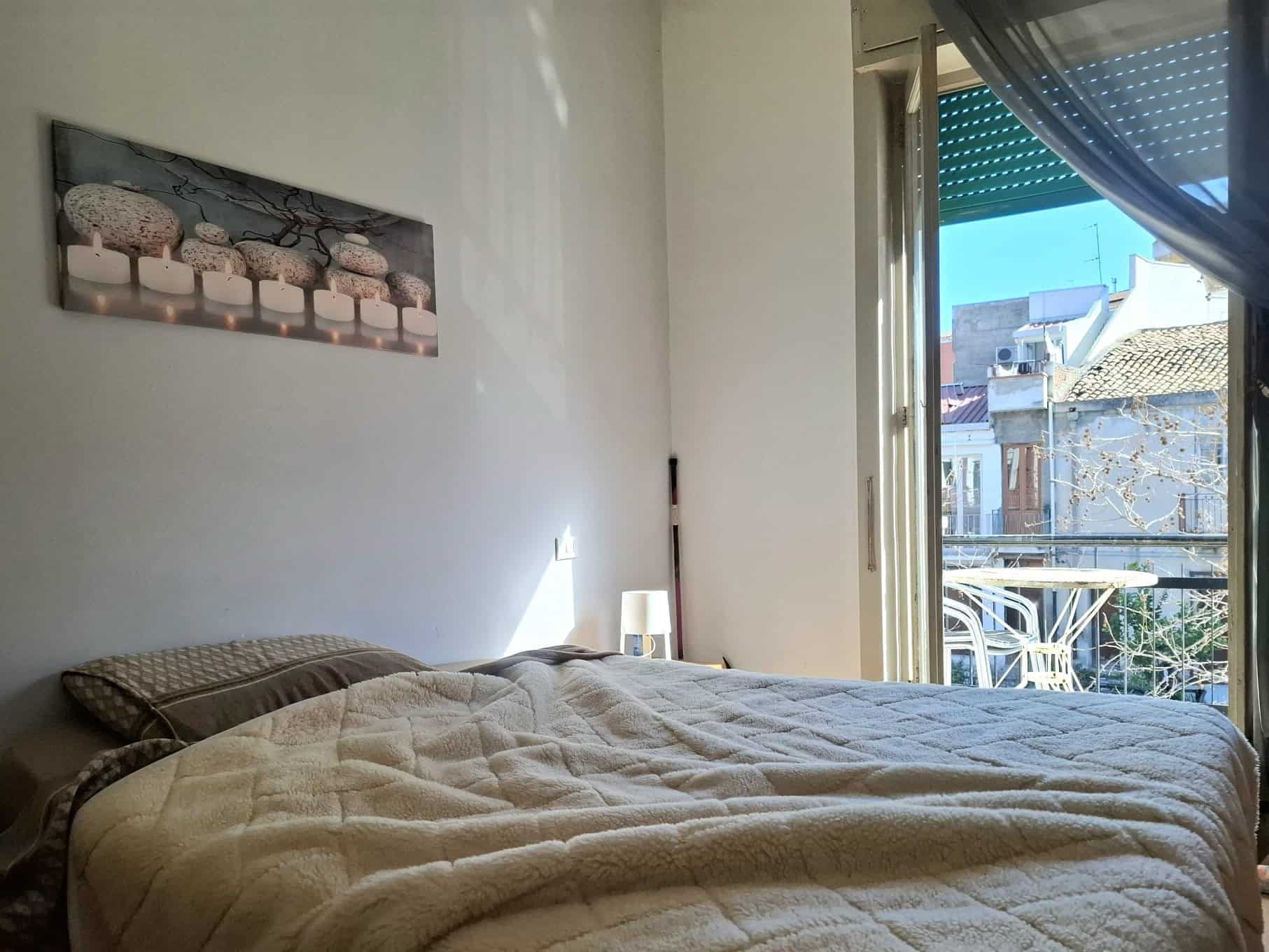 Camera da letto luminosa con letto matrimoniale, vista su balcone e arredamento semplice, ambiente accogliente e soleggiato.