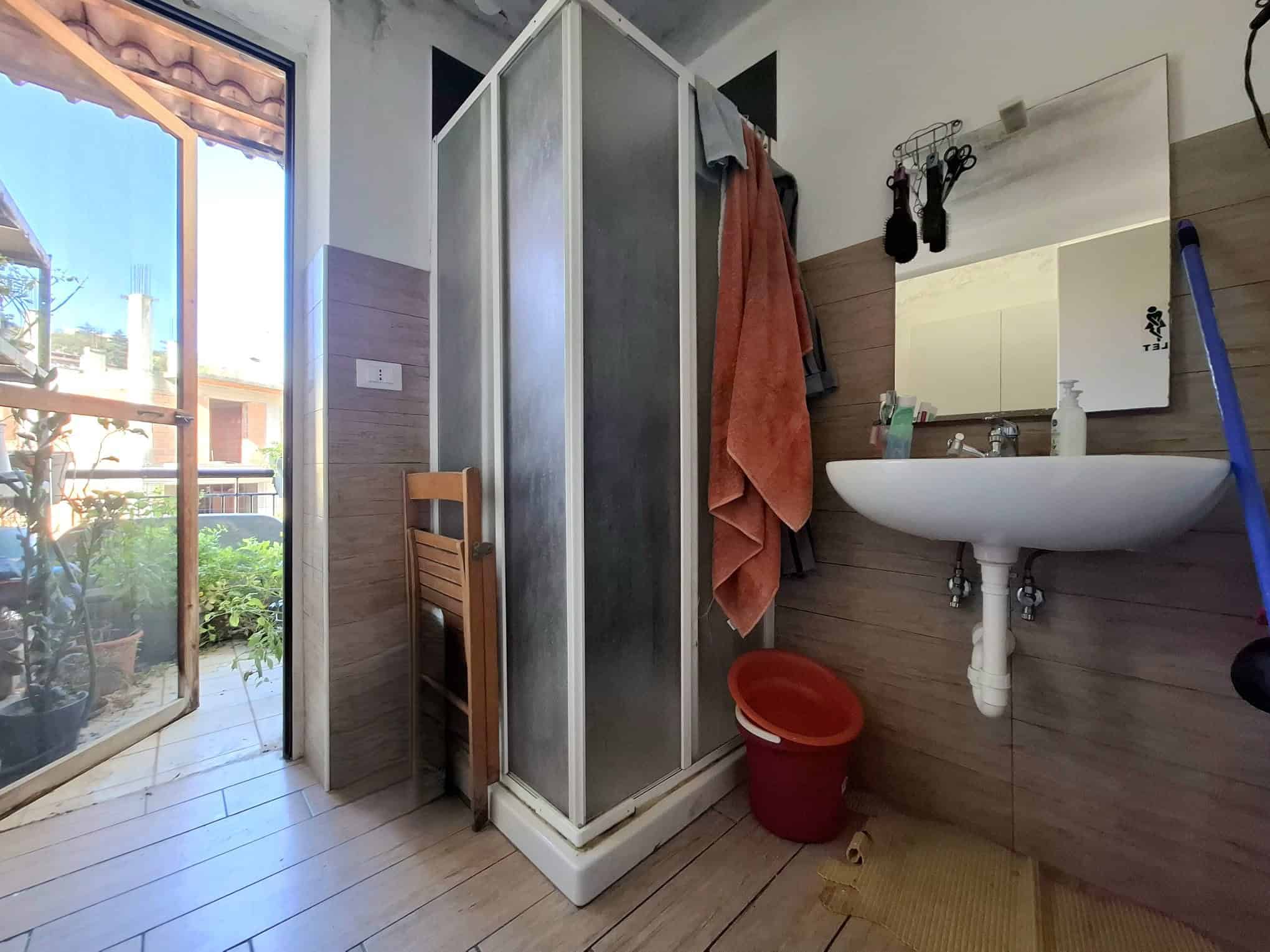 Doccia in cabina con pareti in vetro e accessori per il bagno, illuminata dalla luce naturale proveniente dalla porta finestra che dà su un balcone con piante e case circostanti.