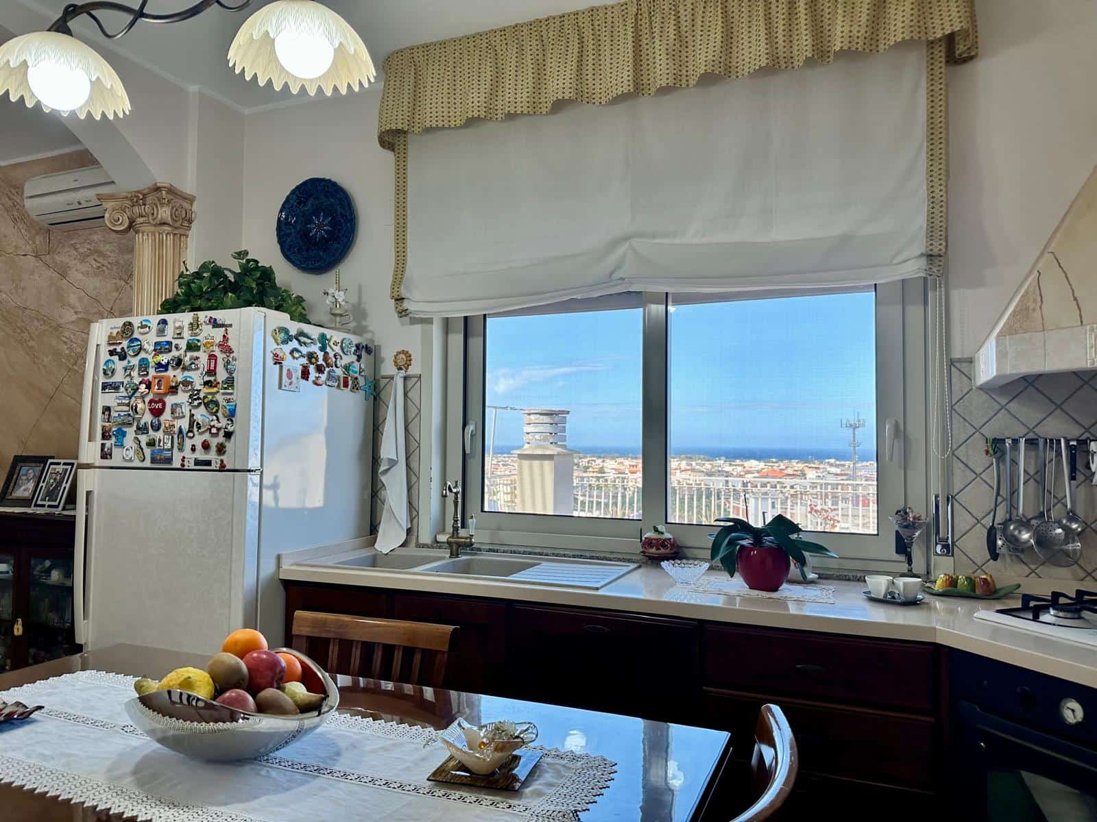 Cucina moderna con vista panoramica sulla città, frigorifero decorato con magneti, e dettagli eleganti, ambiente luminoso e accogliente.