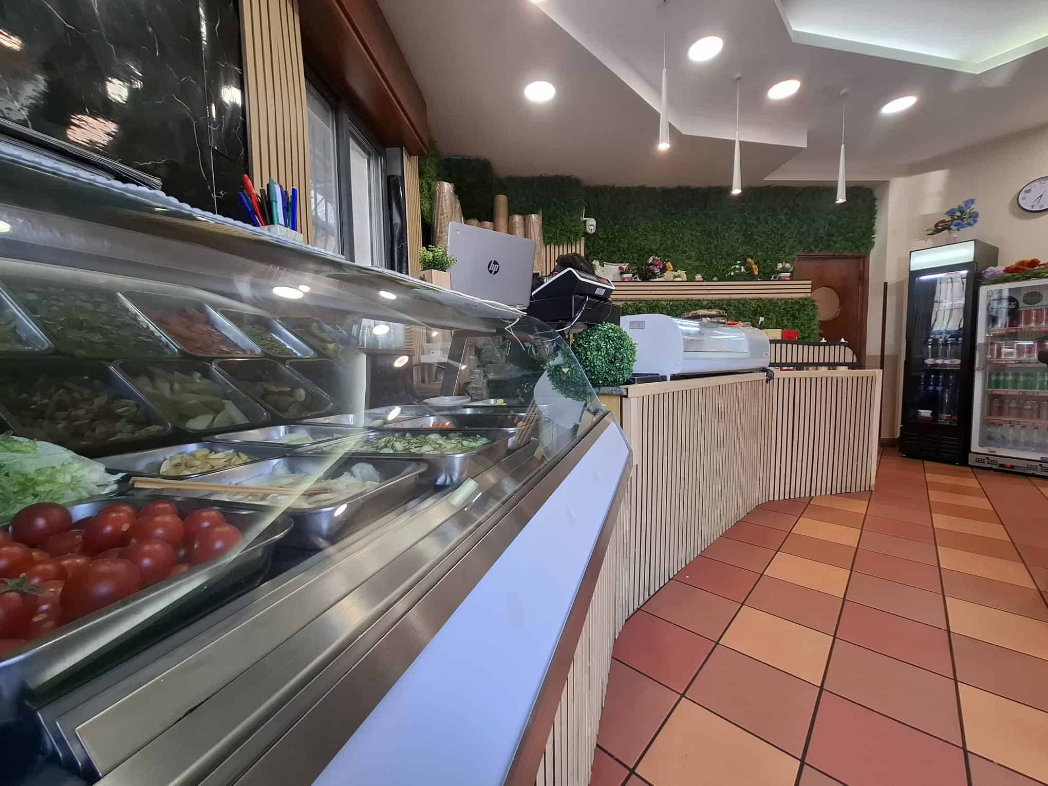 Piatto di insalate fresche e ingredienti biologici in un banco refrigerato di un ristorante o negozio di alimentari, ambiente moderno e accogliente con decorazioni verdi e pavimento in piastrelle.