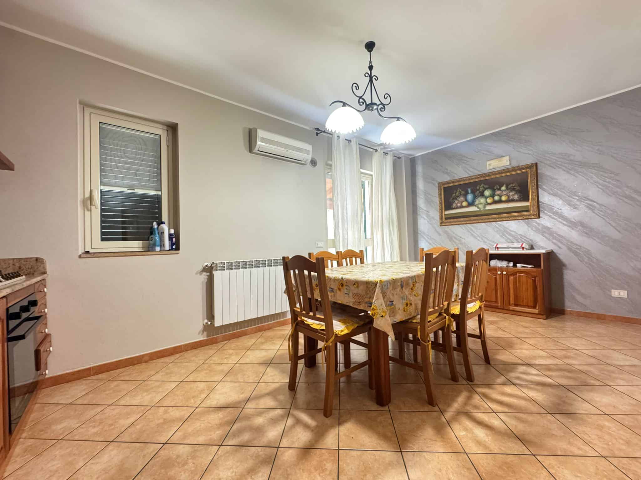 Ampia sala da pranzo con tavolo in legno e sedie, illuminata da lampadario centrale, con pareti dipinte in tonalità neutre e decorazioni floreali, ideale per convivialità e cene in famiglia.