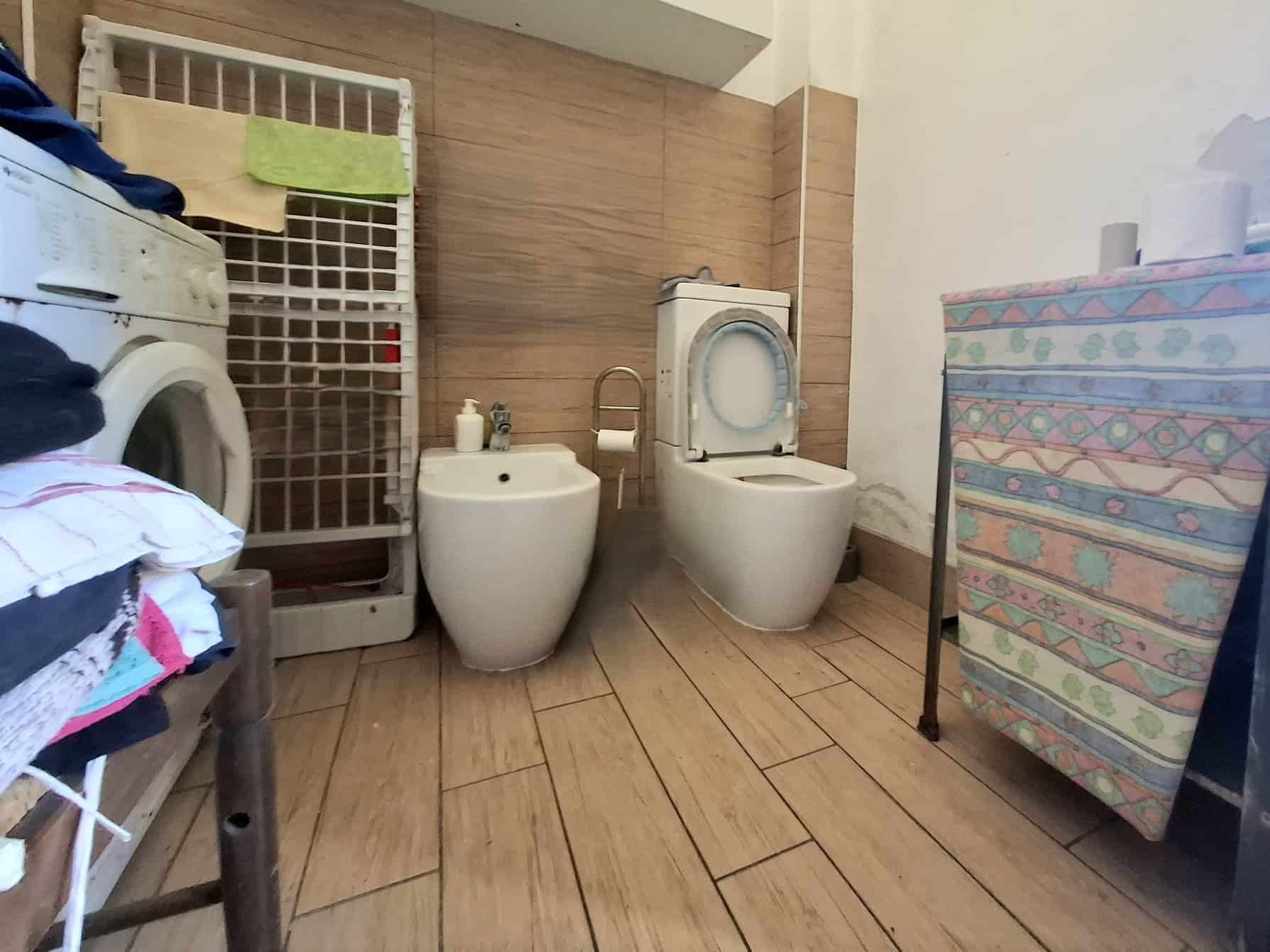 Lavanderia con lavatrice, bidet e WC in un ambiente domestico con pavimento in piastrelle e pareti rivestite in legno. Spazio funzionale e pratico per le faccende quotidiane.