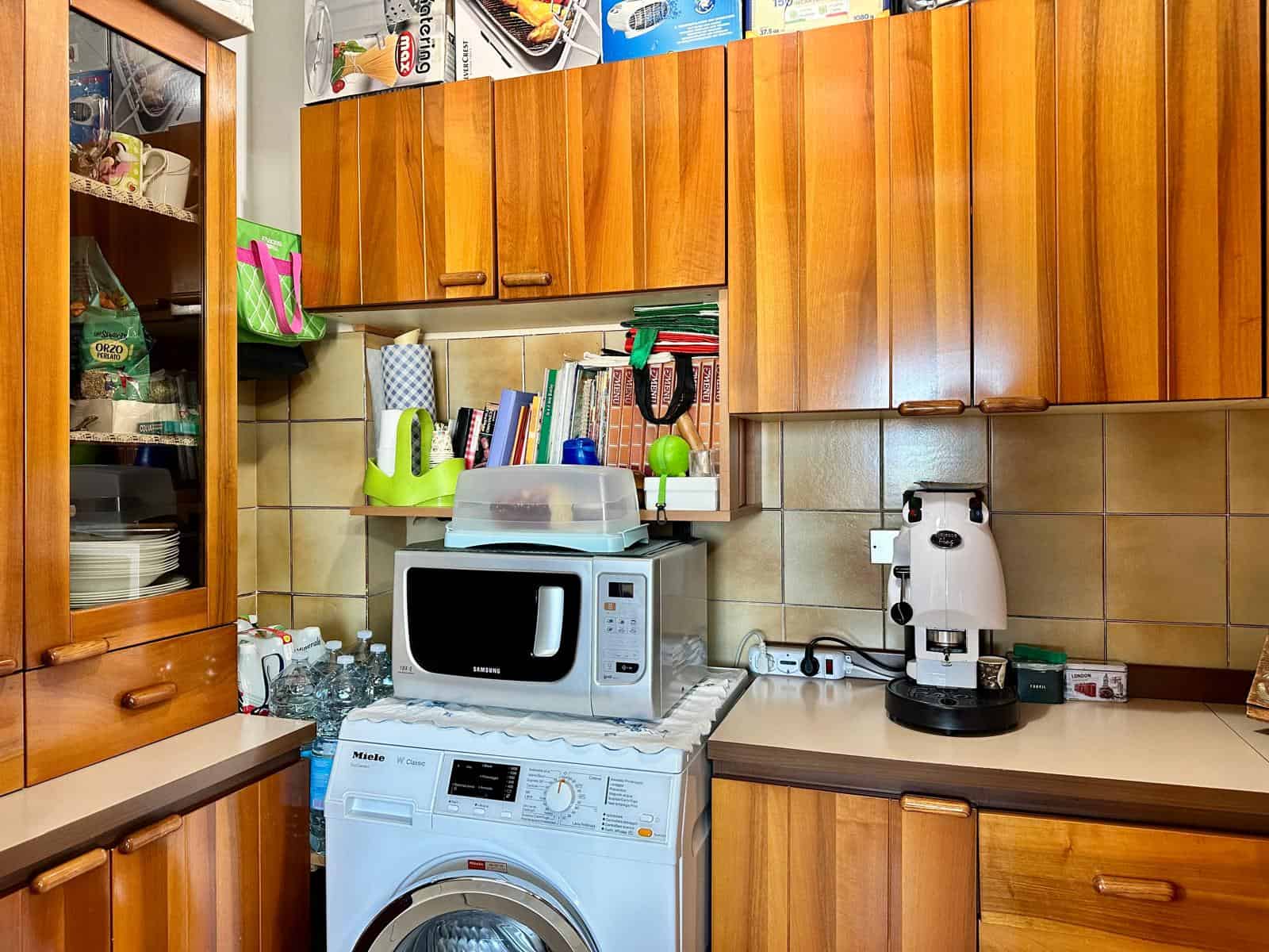 Pannello di cucina con armadietti in legno, microonde, lavatrice e macchina da caffè, ambiente domestico funzionale e ben organizzato.