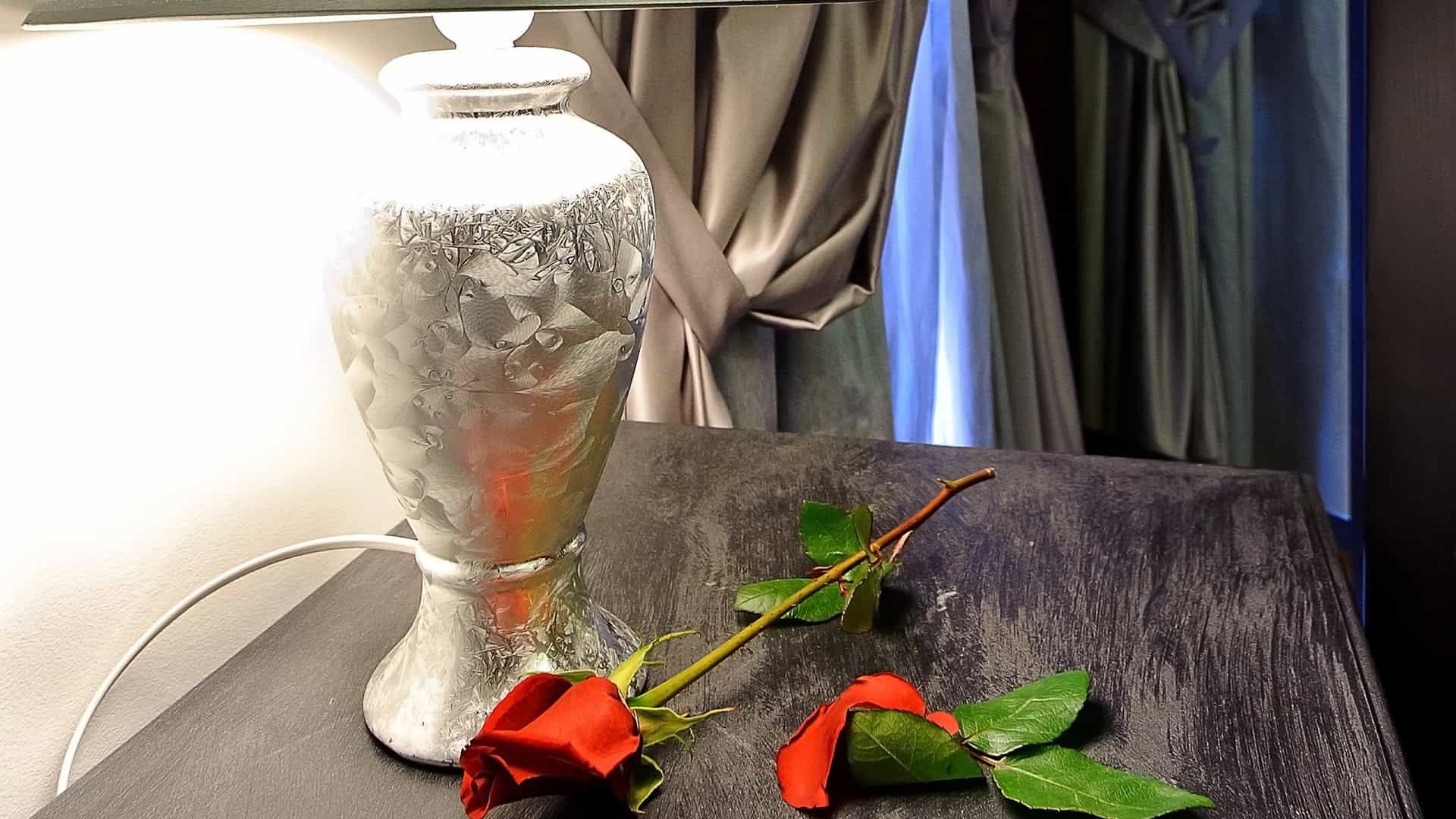 Elegante lampada da tavolo in vetro con riflessi, decorata con rose rosse fresche e foglie verdi, su un tavolo in legno scuro in un ambiente raffinato e accogliente.