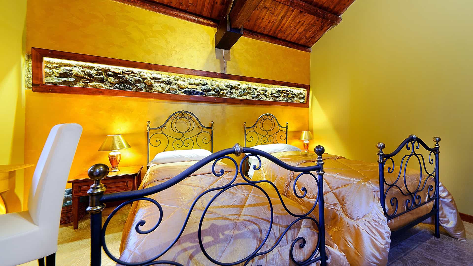 Letto matrimoniale con testiera in ferro battuto in una camera accogliente con pareti gialle e soffitto in legno. Atmosfera calda e rilassante, ideale per soggiorni confortevoli e romantici.