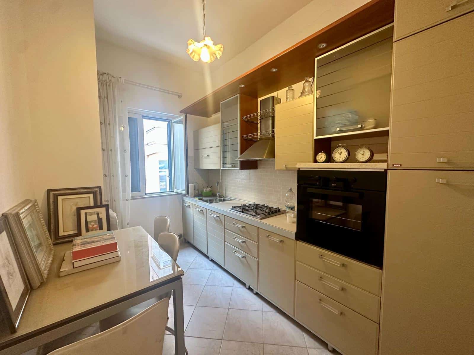 Cucina moderna con mobili chiari, piano di lavoro in marmo e elettrodomestici integrati, illuminata da luce naturale e lampada a soffitto.