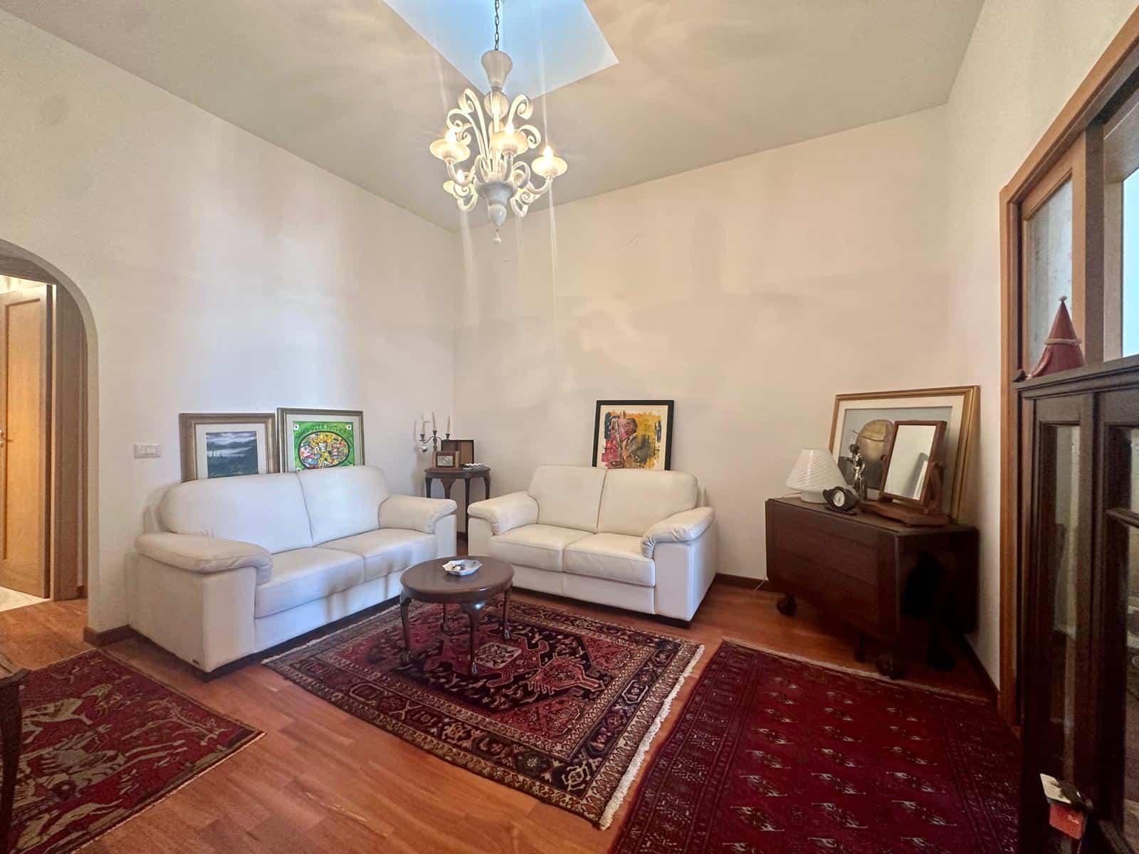 Sedie e divani bianchi in soggiorno con tappeti rossi e lampadario elegante, ambiente accogliente e luminoso.