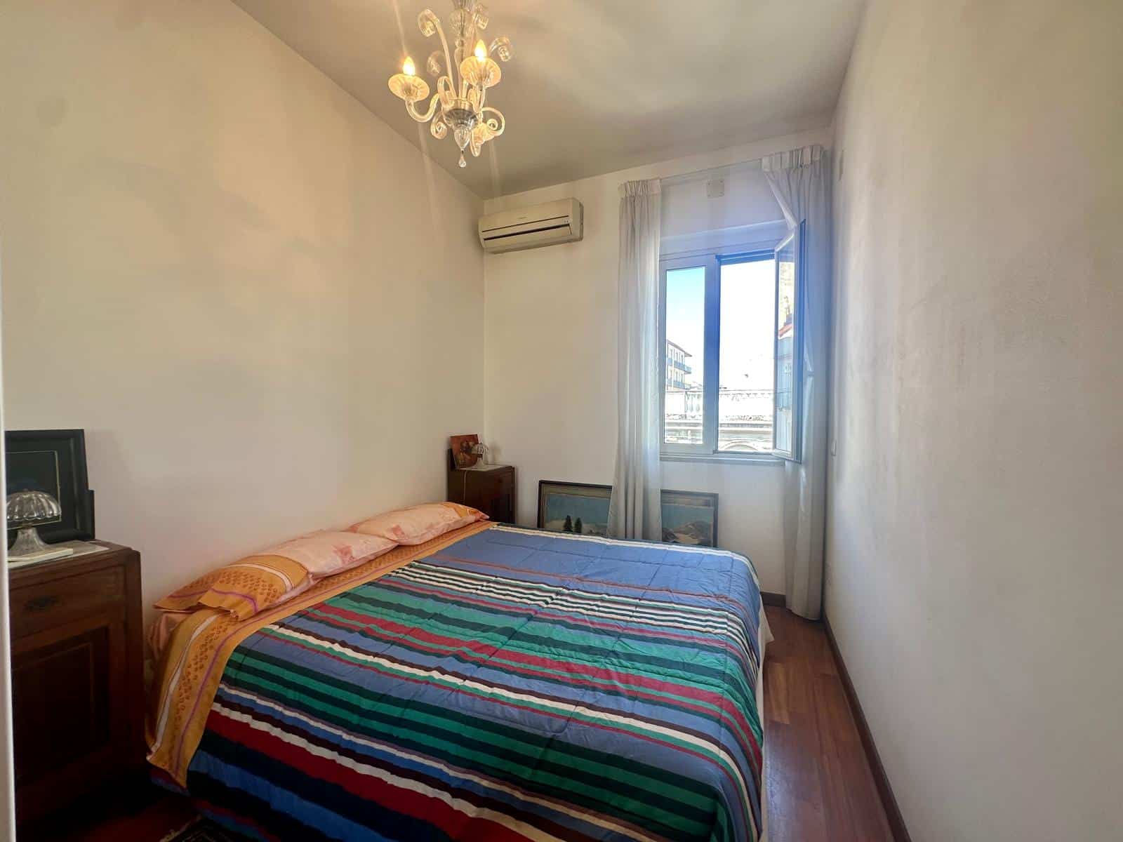 Letto matrimoniale con lenzuola colorate in una stanza luminosa e accogliente, con finestra che permette l'ingresso di luce naturale. Arredamento semplice e funzionale, ideale per ambienti residenzial.