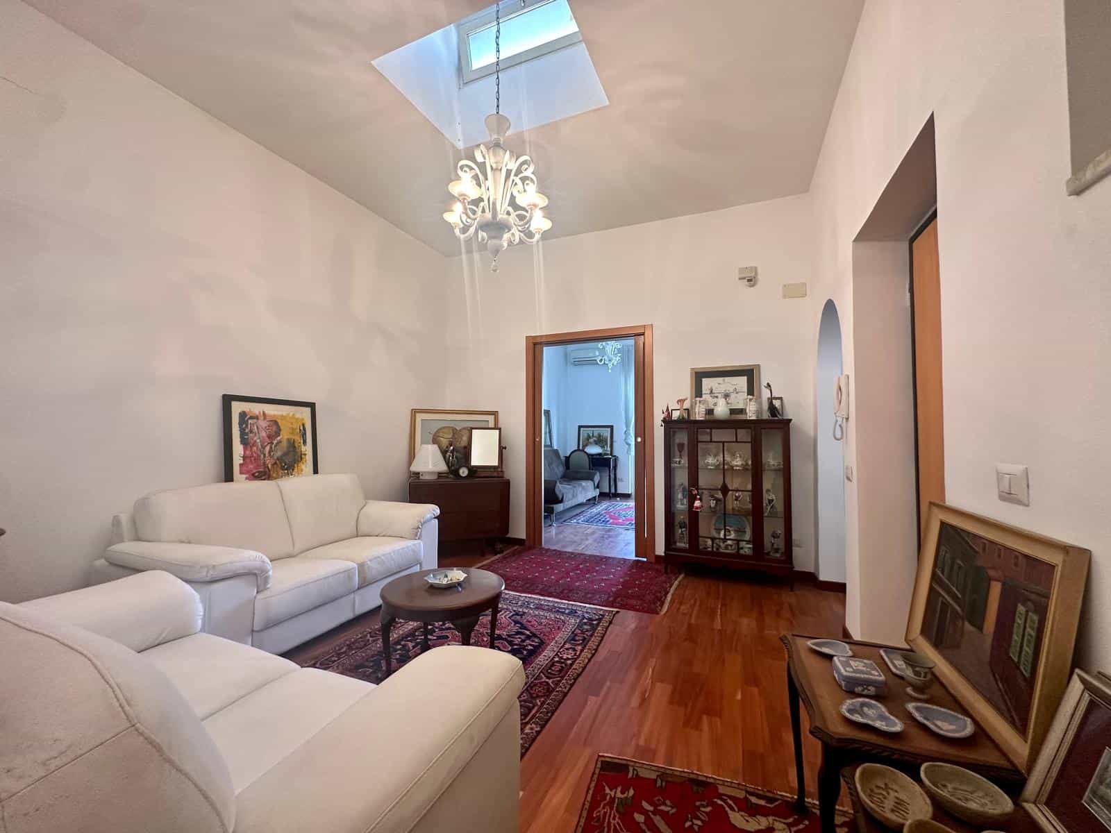 Una luminosa sala da pranzo con soffitto alto, pareti bianche, pavimento in legno e arredi eleganti, tra cui un divano bianco, un tavolino e una credenza in legno, ideale per vivere confortevoli momen.