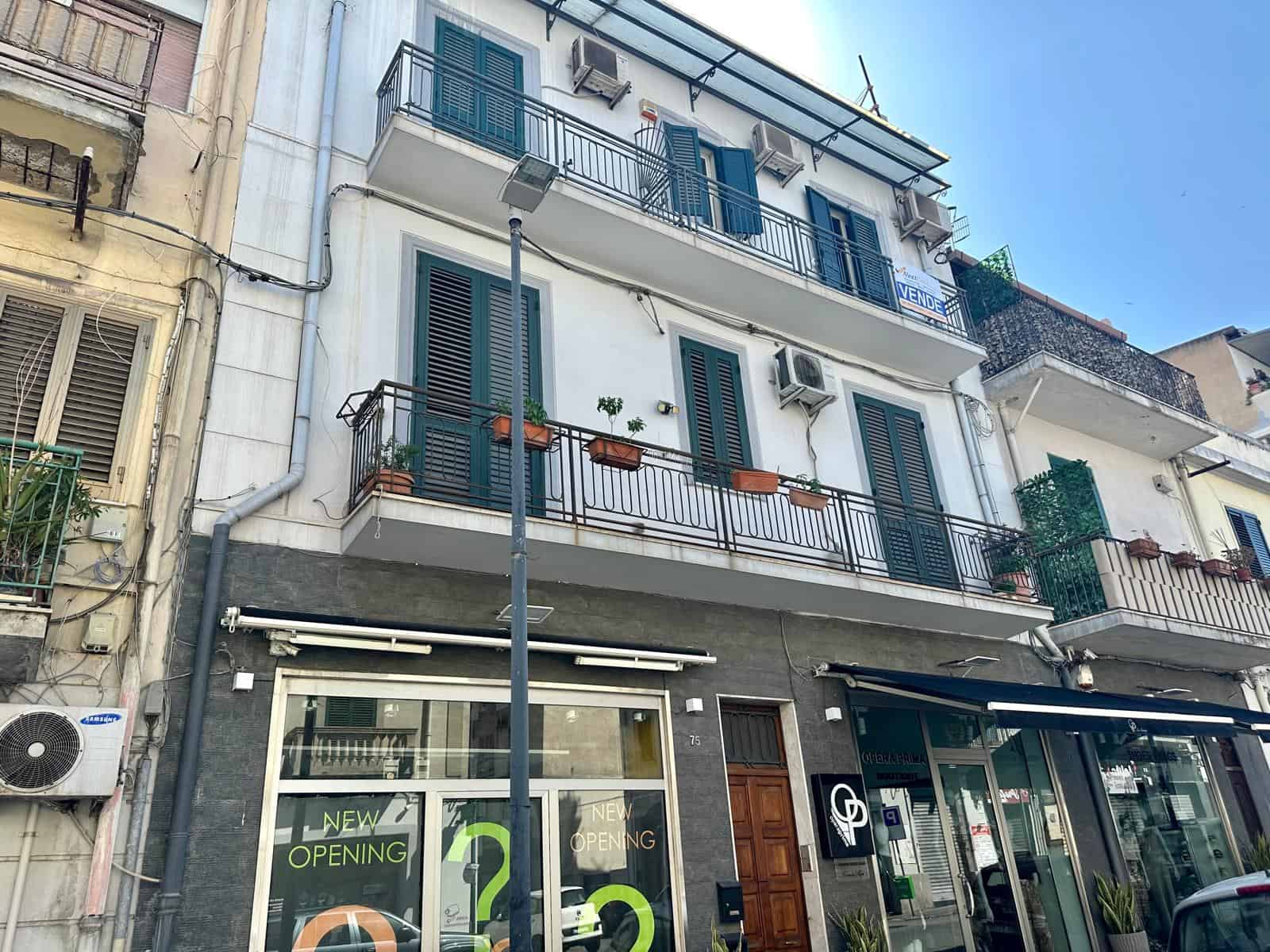 Edificio residenziale con balconi e negozio al piano terra, in zona urbana, con dettagli architettonici moderni e piante sui balconi.
