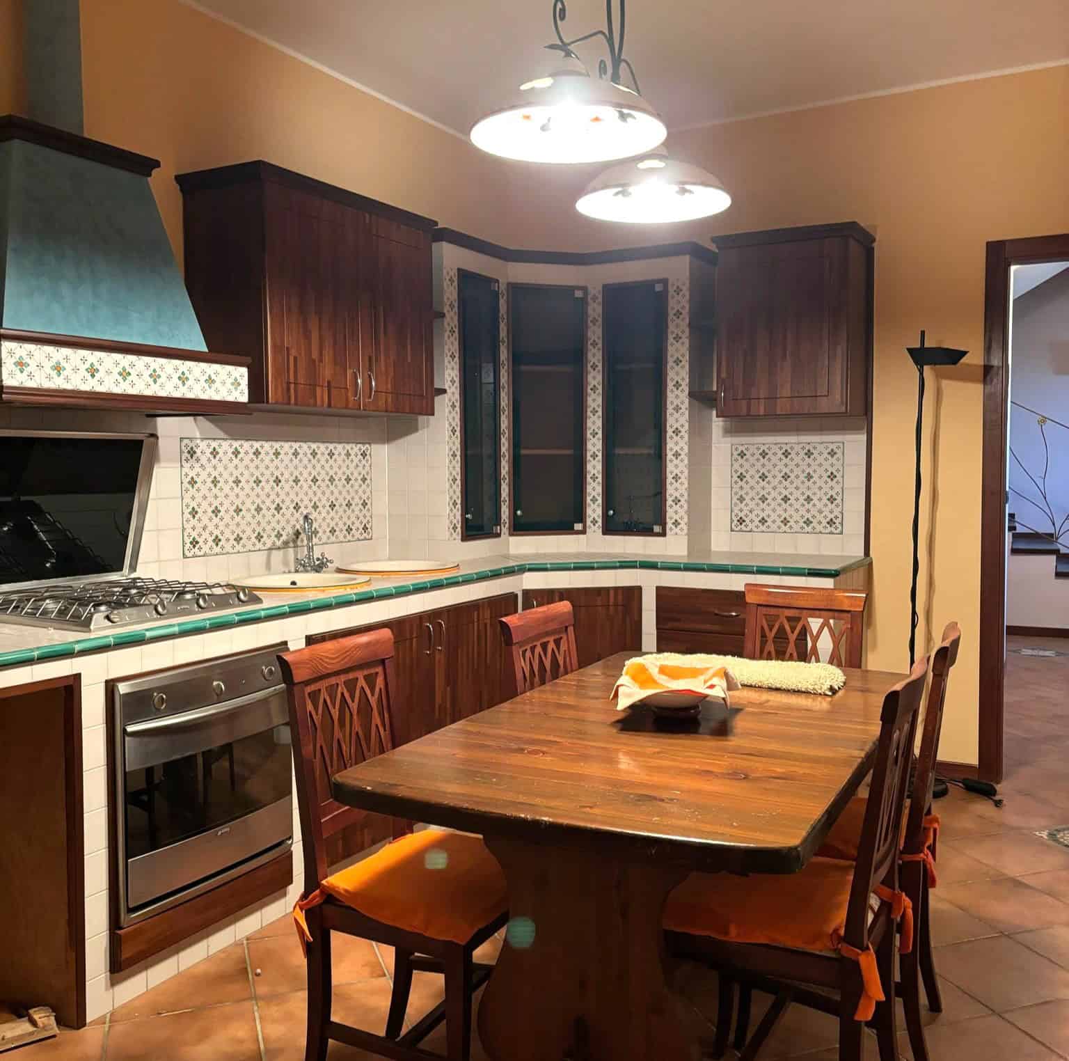 Piano cucina in stile classico con mobili in legno scuro, piano di lavoro in ceramica e pareti decorate con piastrelle colorate, illuminato da lampadari moderni.