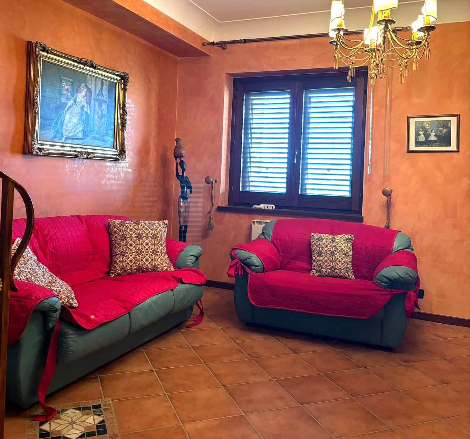 Soggiorno accogliente con divani verdi e coperte rosse, pareti arancioni e decorazioni classiche, tra cui quadri e lampadario in stile antico, illuminato da una finestra con persiane blu.
