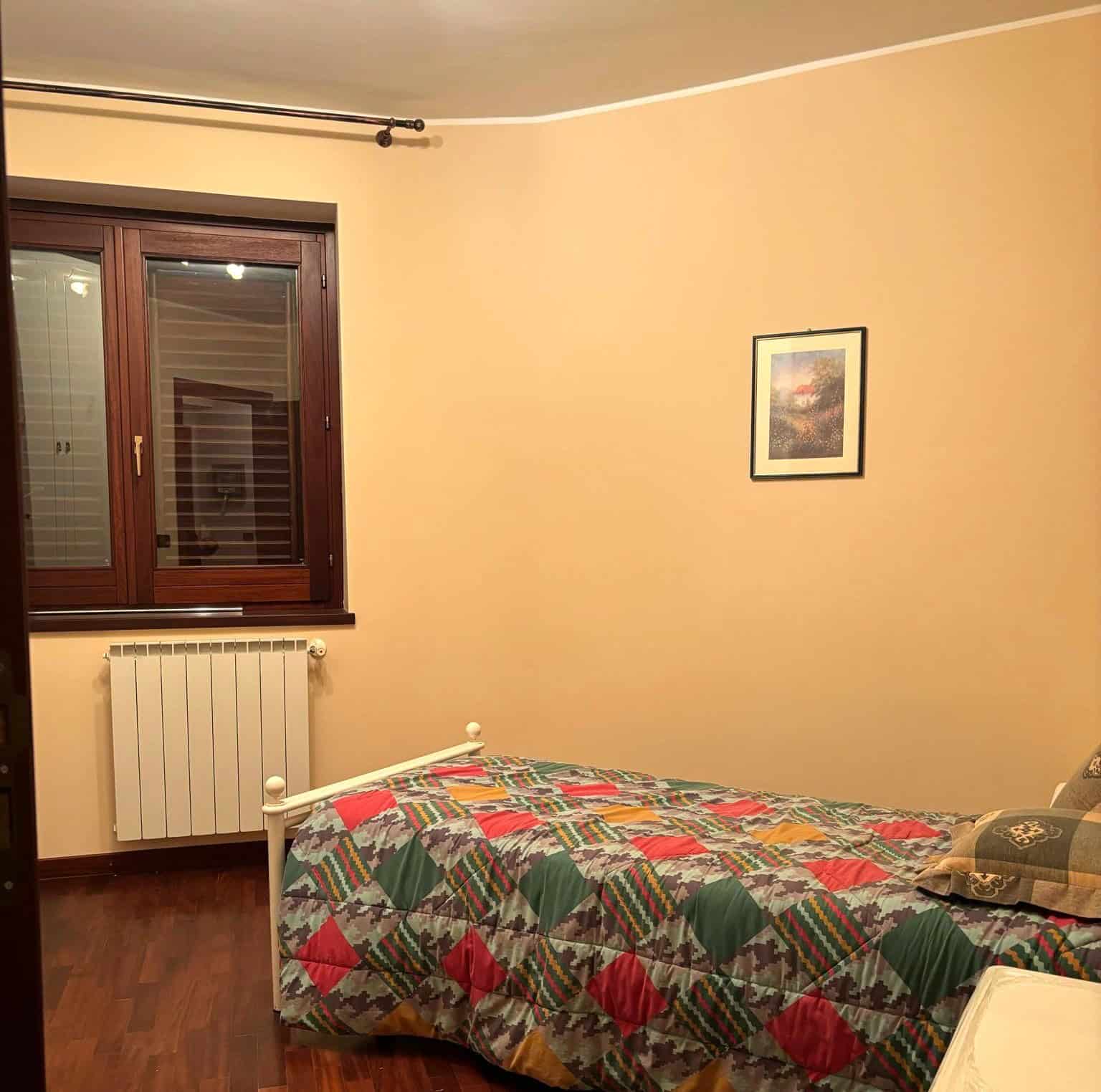 Camera da letto con pareti beige, finestra con persiane e letto singolo con copriletto colorato, ambiente accogliente e luminoso.
