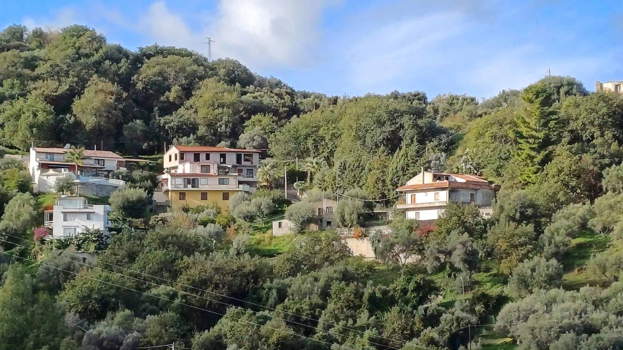 Vista panoramica di case indipendenti in vendita in una zona collinare di Capo d'Orlando, con vegetazione lussureggiante e cielo sereno.