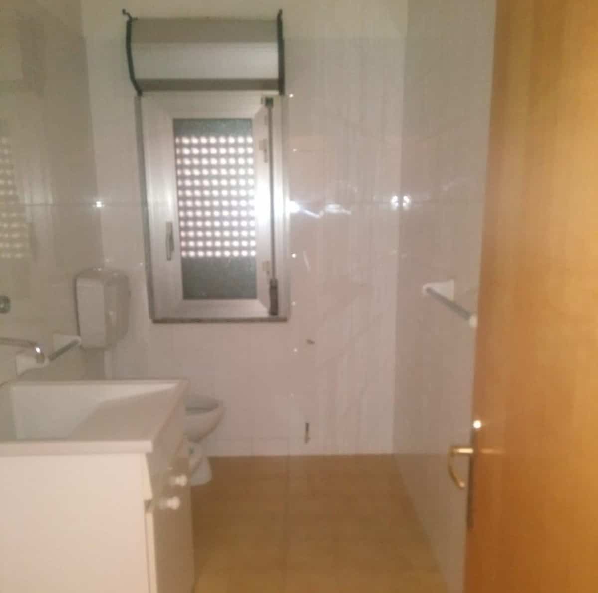 Lavabo e servizi igienici in un bagno di piccole dimensioni con pareti rivestite in piastrelle chiare, finestra con tapparella e pavimento in gres beige, ambiente funzionale e semplice.