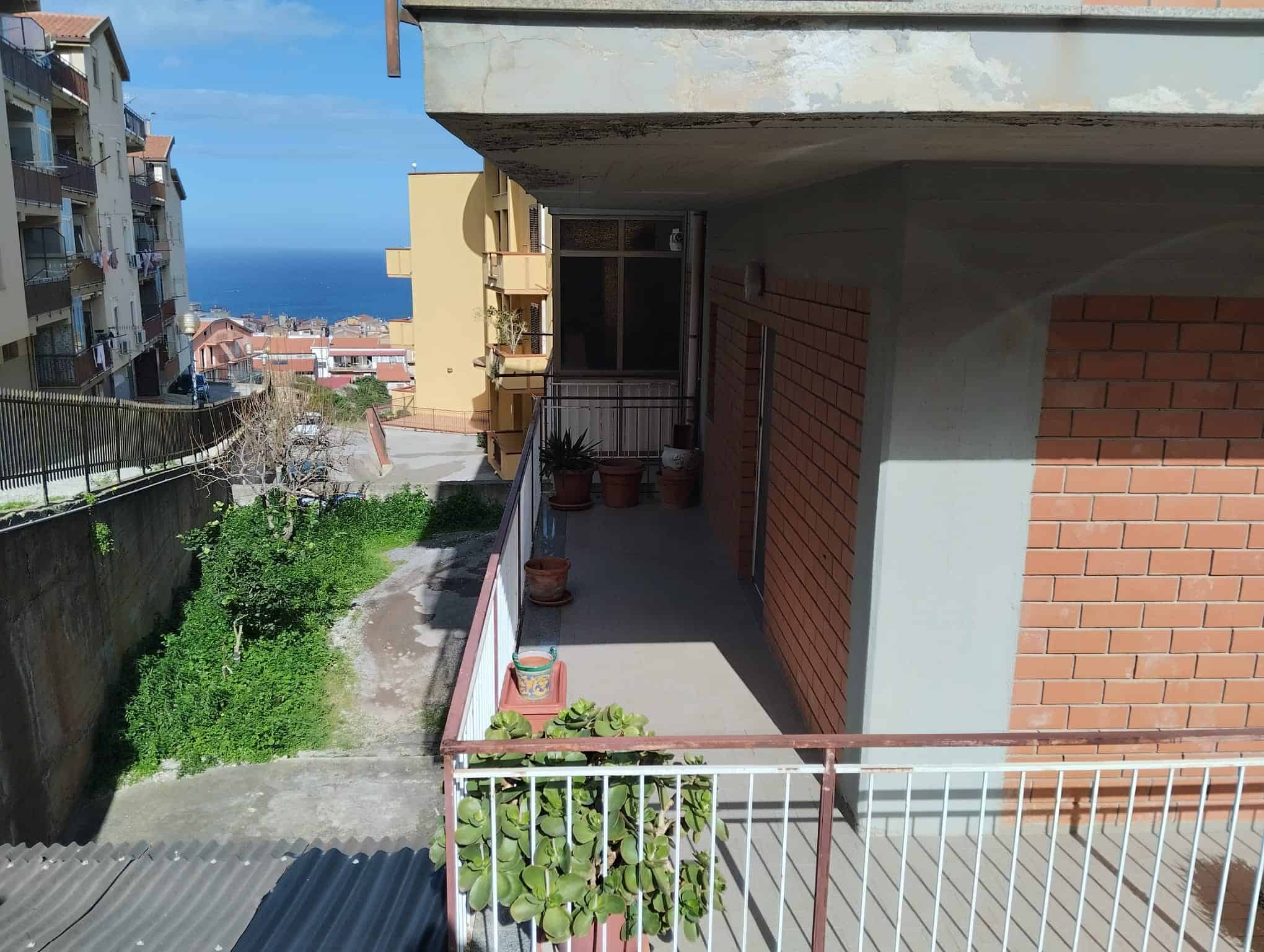 Balcone di un appartamento con vista mare, con piante in vaso e ringhiera in metallo, in un contesto urbano residenziale, con edifici circostanti e il mare sullo sfondo, in una giornata soleggiata.