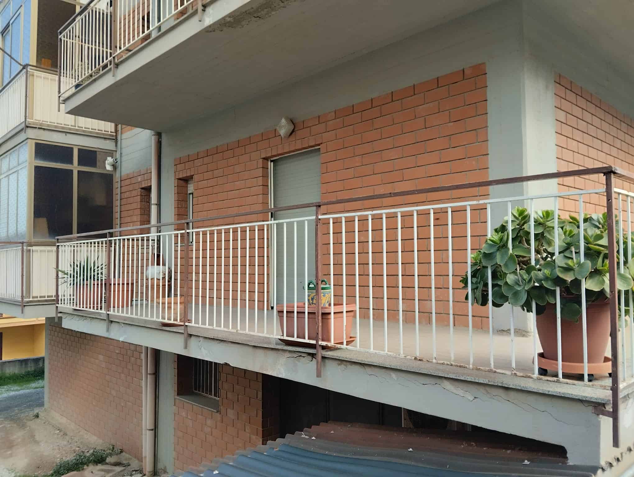 Porte di accesso a un appartamento con balcone in cemento e ringhiera in metallo, decorato con vasi di piante verdi, in un edificio residenziale in mattoni rossi.