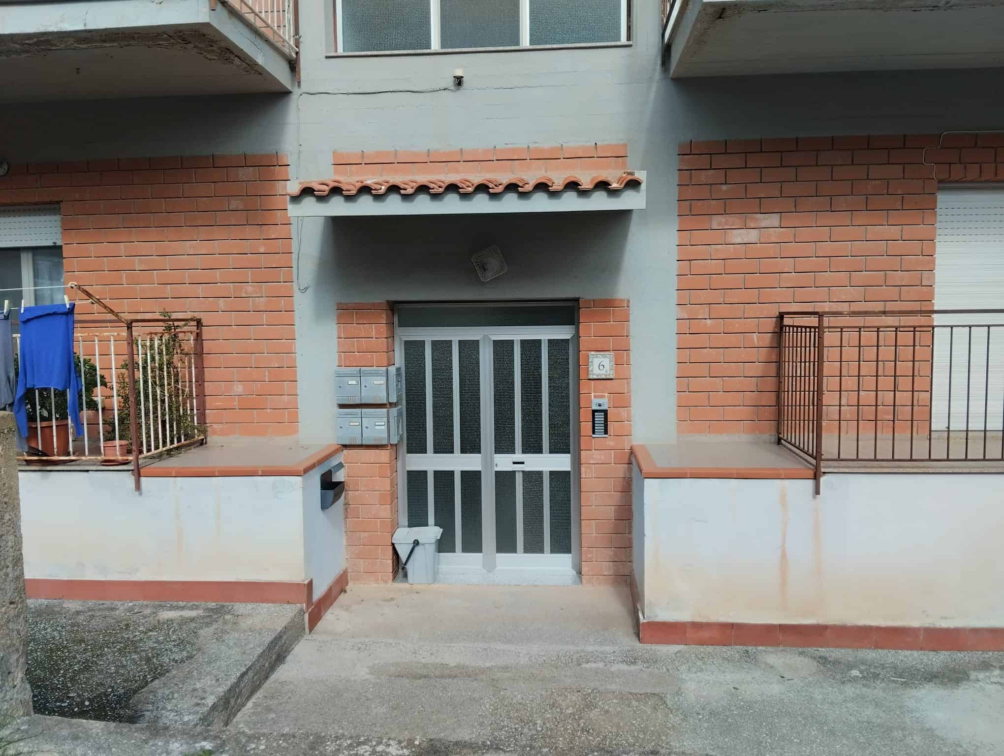 Portone d'ingresso in metallo con grate, situato tra due pareti in mattoni rossi e rivestimenti in cemento, con accesso pedonale e area di ingresso pavimentata.