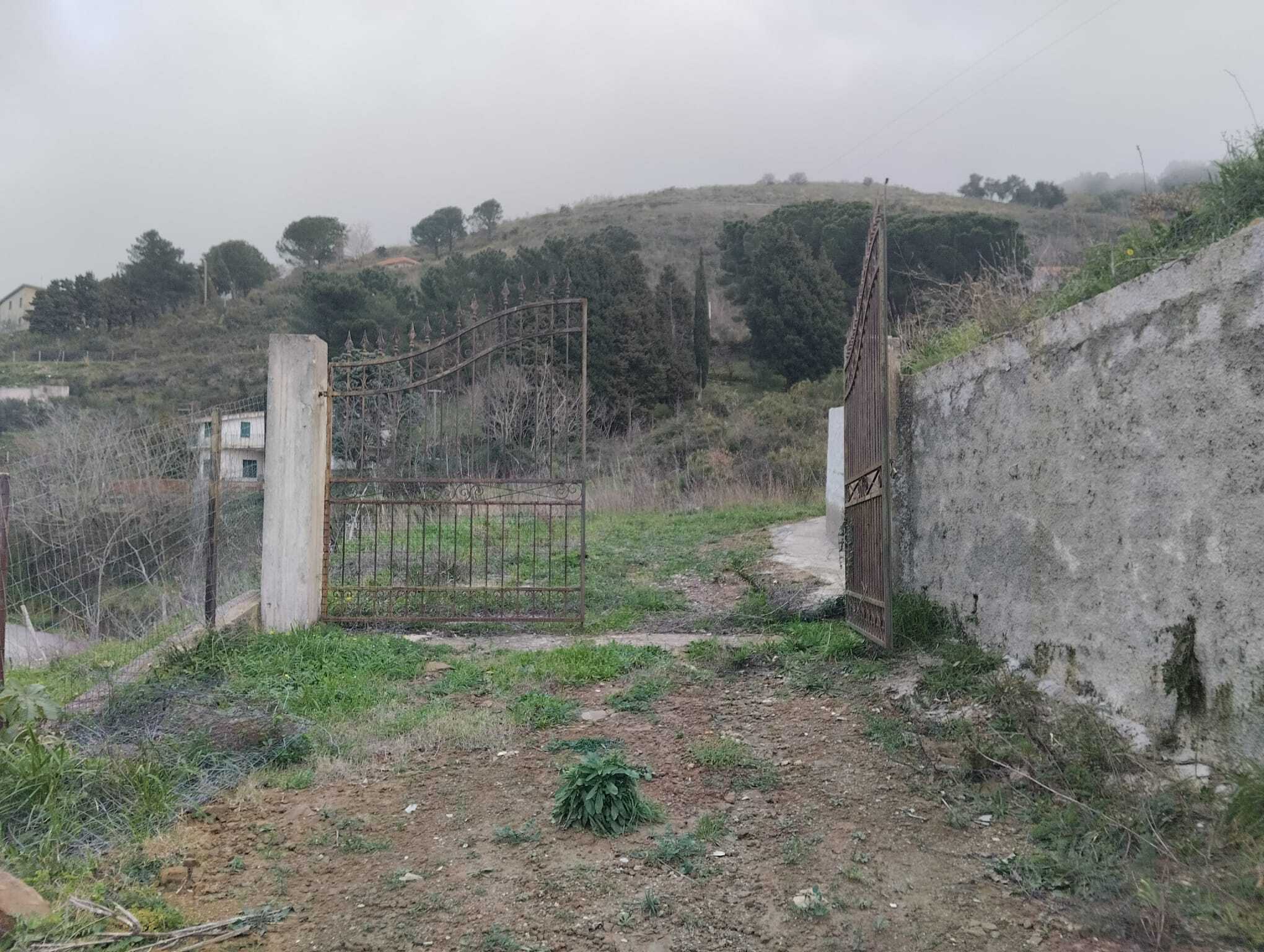 Cancello rustico in ferro battuto aperto su un sentiero di campagna con paesaggio collinare e vegetazione naturale, ideale per proprietà rurali o terreni agricoli.