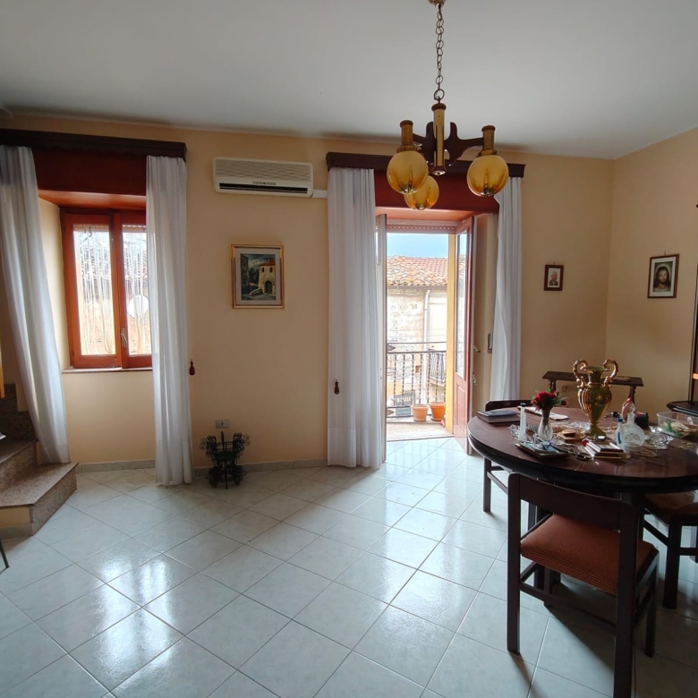 Interno soggiorno luminoso con grandi finestre e accesso al balcone, arredato con tavolo e sedie, decorazioni classiche e aria condizionata, ideale per vivere in comfort.