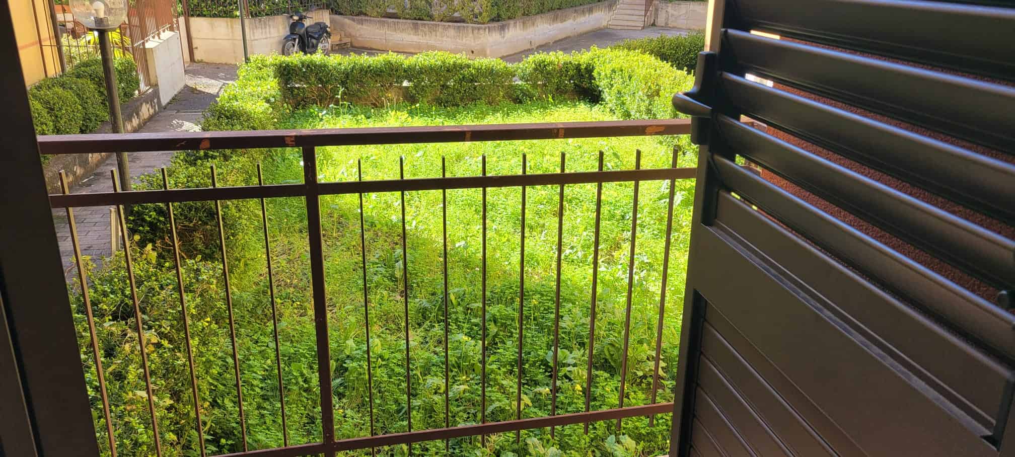 Immagine di un balcone con ringhiera in metallo che si affaccia su un giardino verde e curato, con piante e siepi ben mantenute.
