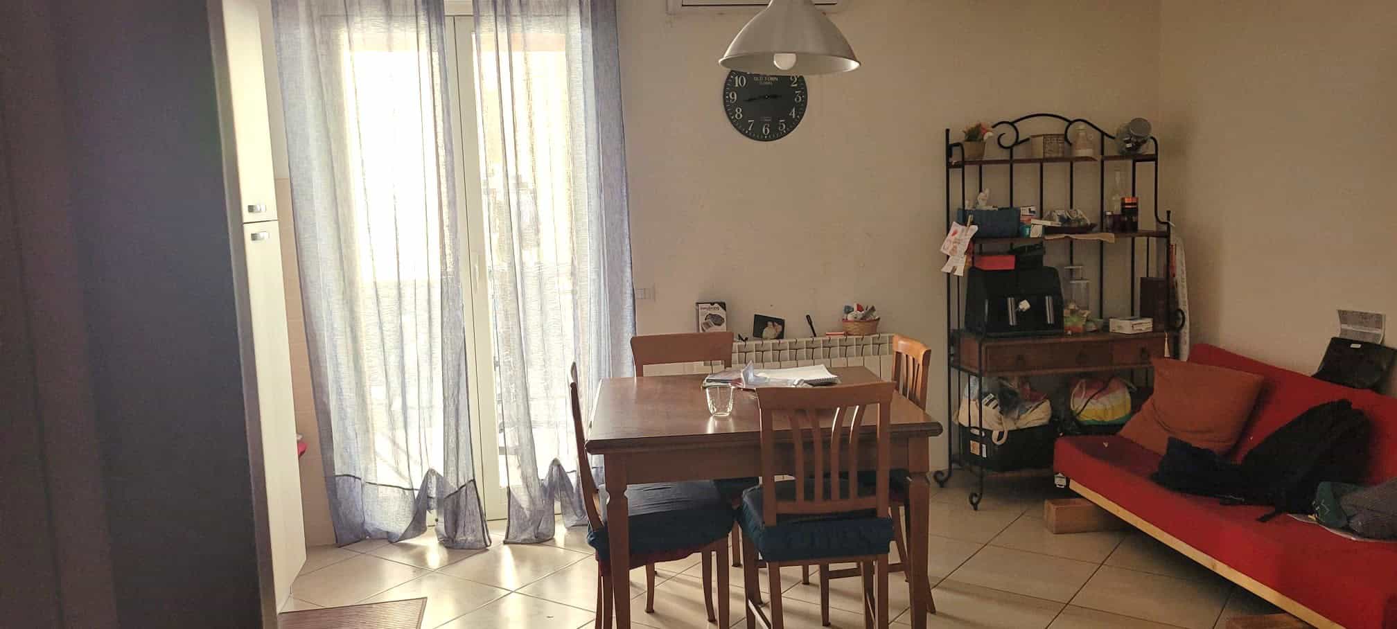Ampia sala da pranzo con tavolo in legno e sedie, illuminata da luce naturale attraverso grandi finestre con tende leggere, arredamento funzionale e accogliente.